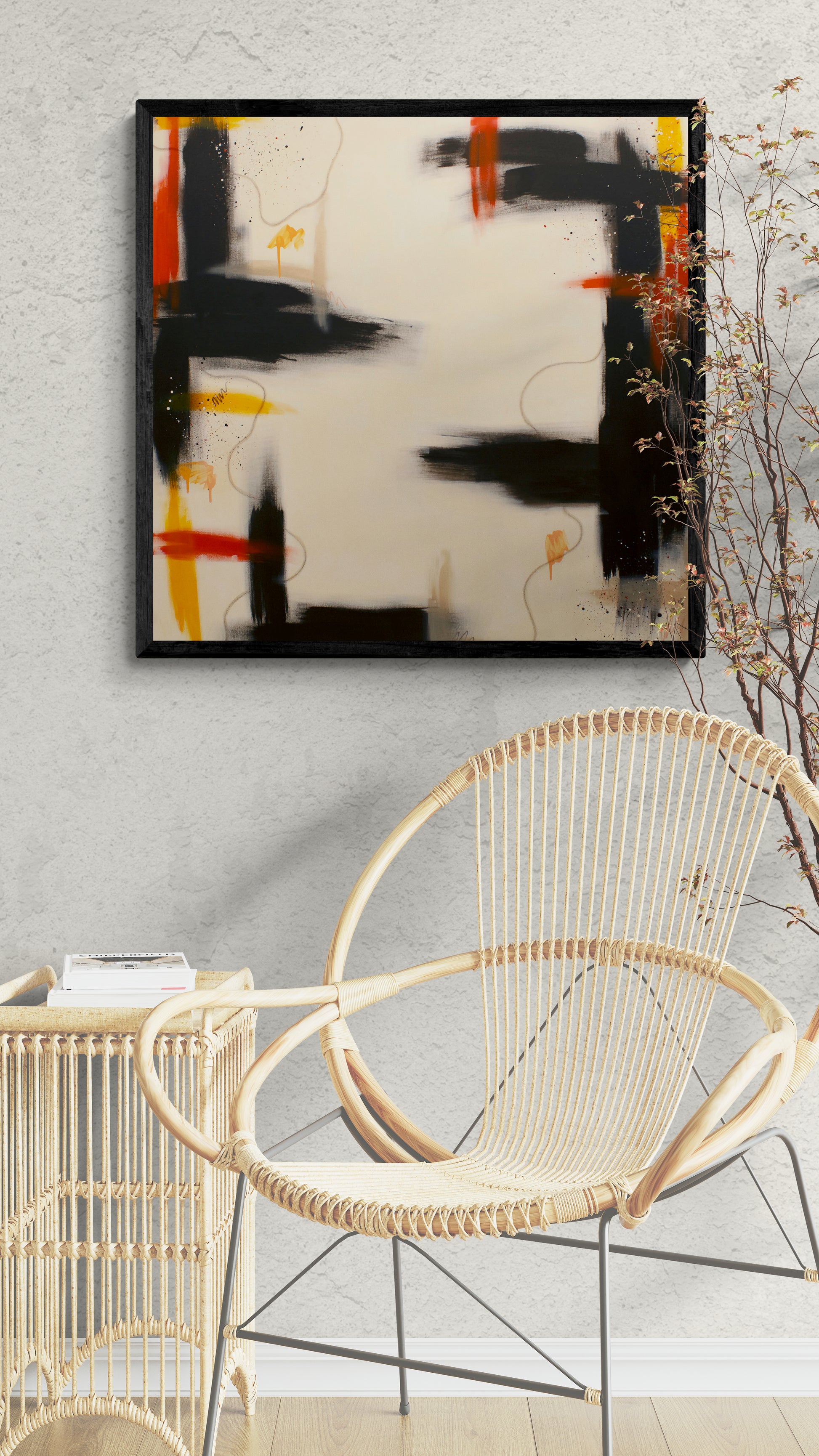 A square contemporary abstract canvas print in a black frame, featuring bold brown brush strokes with vibrant orange and orange-yellow accents on a cream background, styled on a light grey textured wall above a rattan chair and side table.