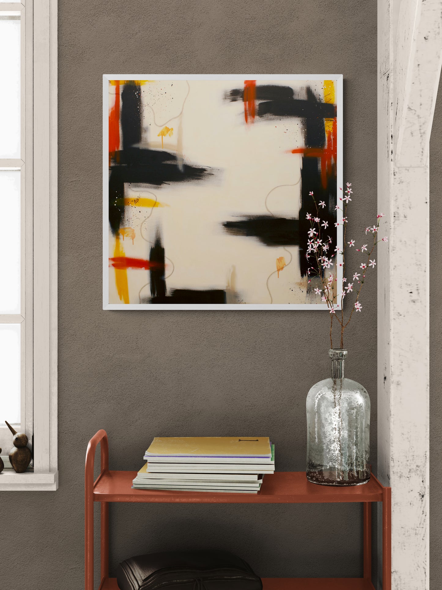A white-framed square abstract art print with sweeping lines and splatters in brown, orange, and orange-yellow, displayed on a dark grey wall above an orange metal console table with books and a glass vase of pink flowers.