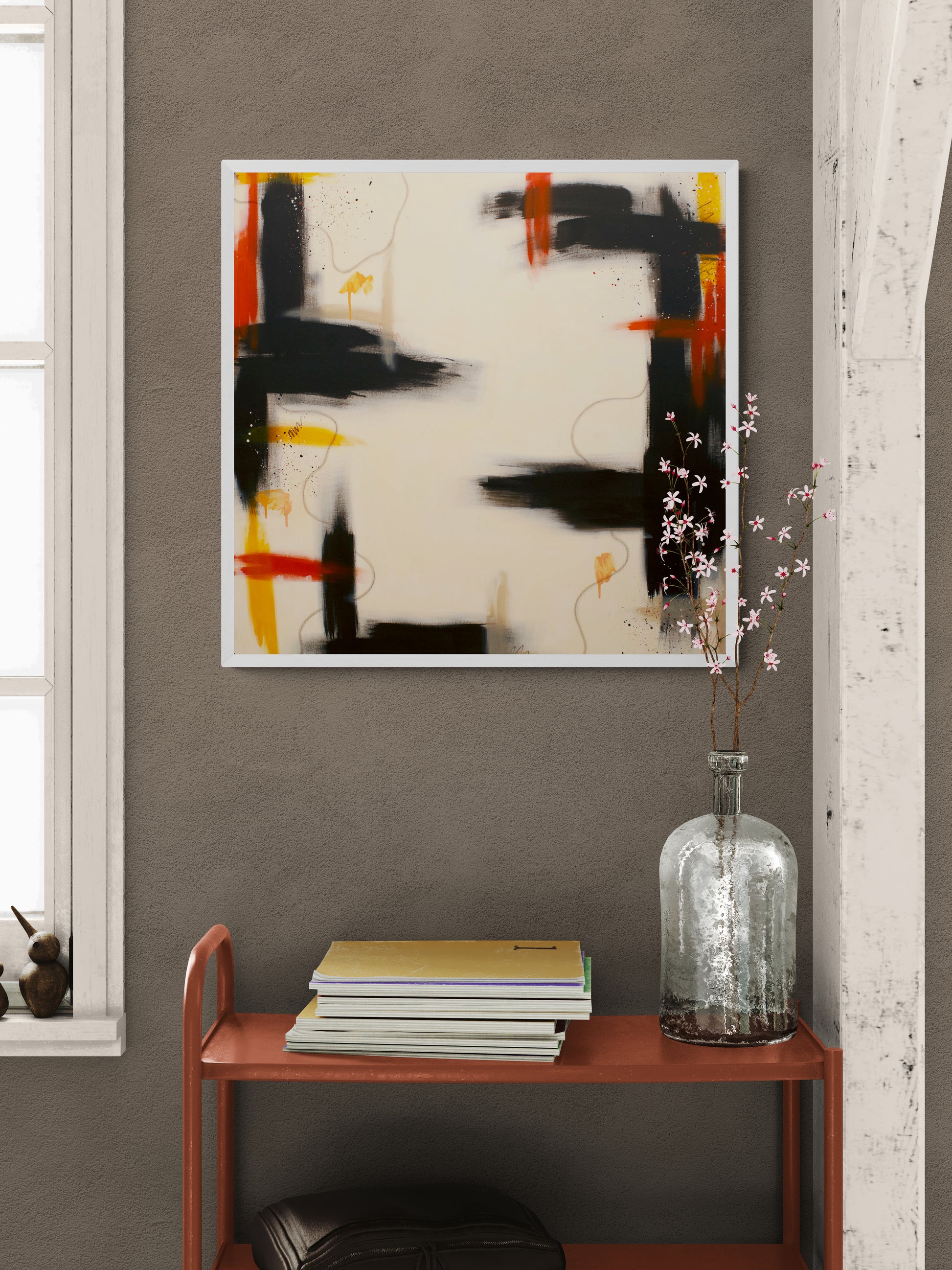 A white-framed square abstract art print with sweeping lines and splatters in brown, orange, and orange-yellow, displayed on a dark grey wall above an orange metal console table with books and a glass vase of pink flowers.