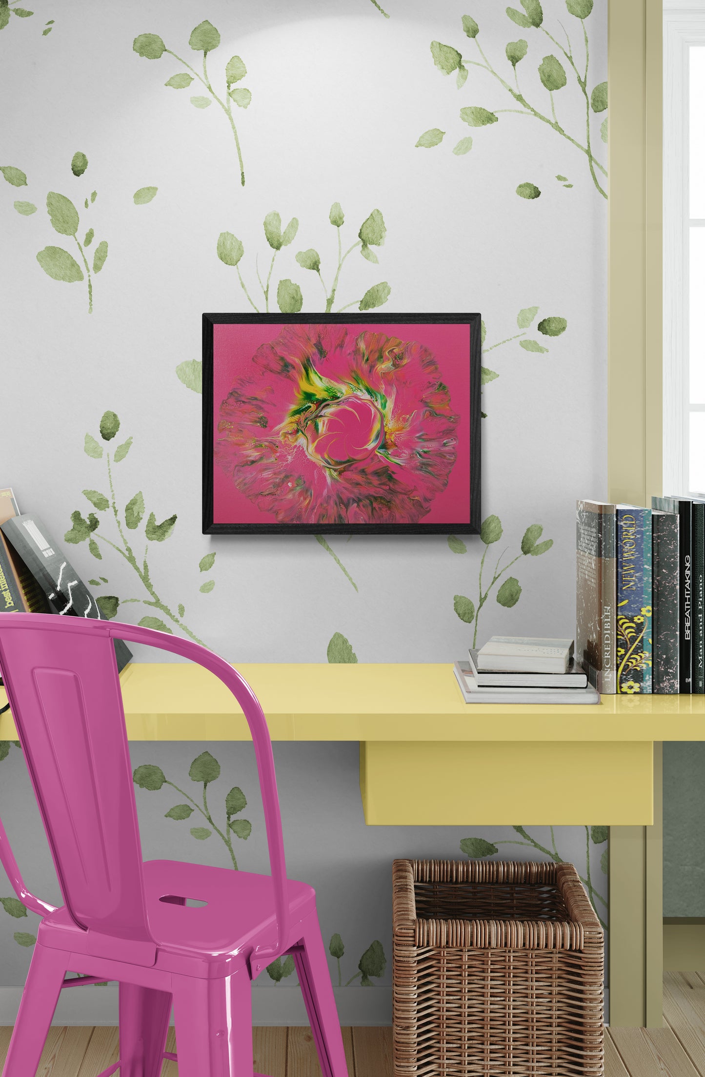 Framed abstract painting with bold pink background and swirling green, yellow, and brown fluid shapes, hung above a yellow desk in a cozy reading nook. The wall behind features green leafy wallpaper, and the setup includes a bright pink metal chair, a basket underneath the desk, and a stack of books on the right side.