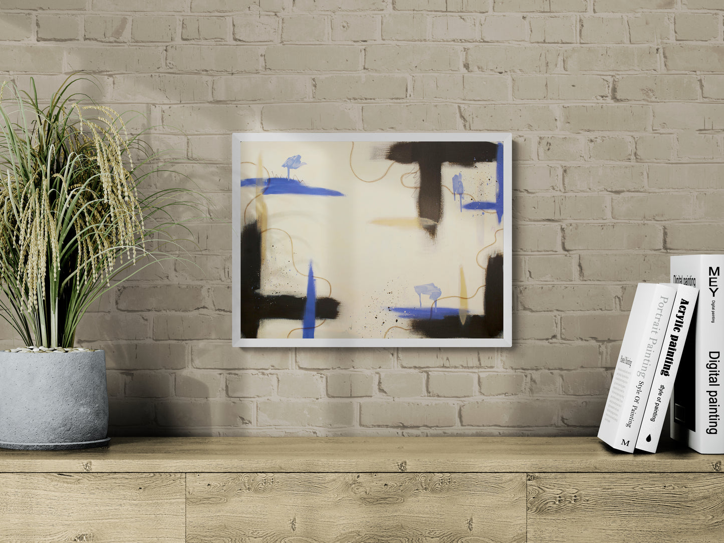 A horizontal abstract painting titled "Suspended Ground" in a white frame, hanging on a light-colored exposed brick wall. The artwork features structured blocks of raw umber contrasted with bright periwinkle blue strokes and thin, wandering gold oil pastel lines on a cream background. To the left is a potted ornamental grass, and to the right are stacked art books on a wooden cabinet.