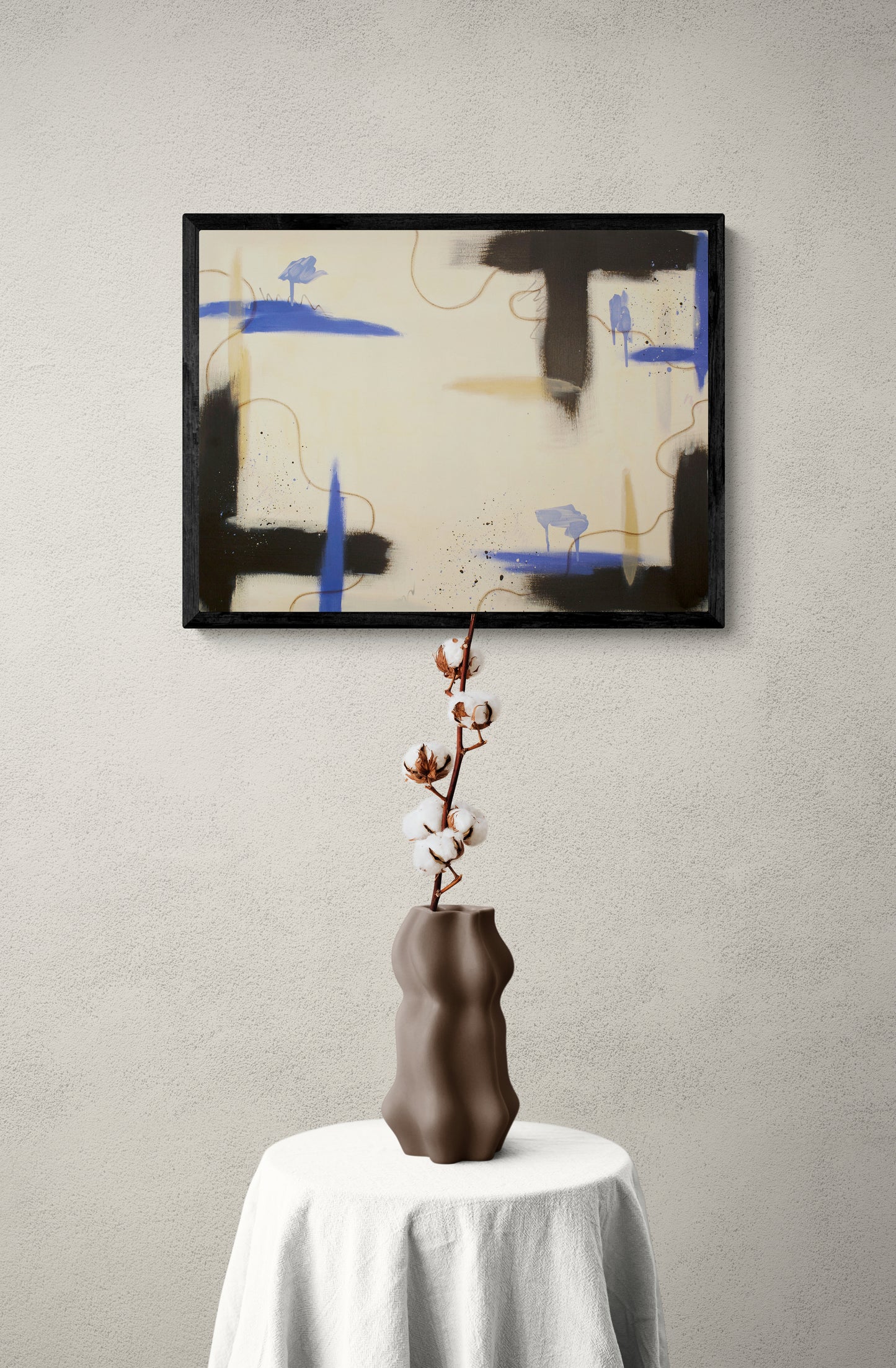 "Suspended Ground" abstract art displayed in a thin black frame on a light beige textured wall. In the foreground, a sculptural brown vase holding a dried cotton branch sits on a round table draped with a white linen tablecloth. The painting’s deep raw umber and blue accents create a modern focal point.