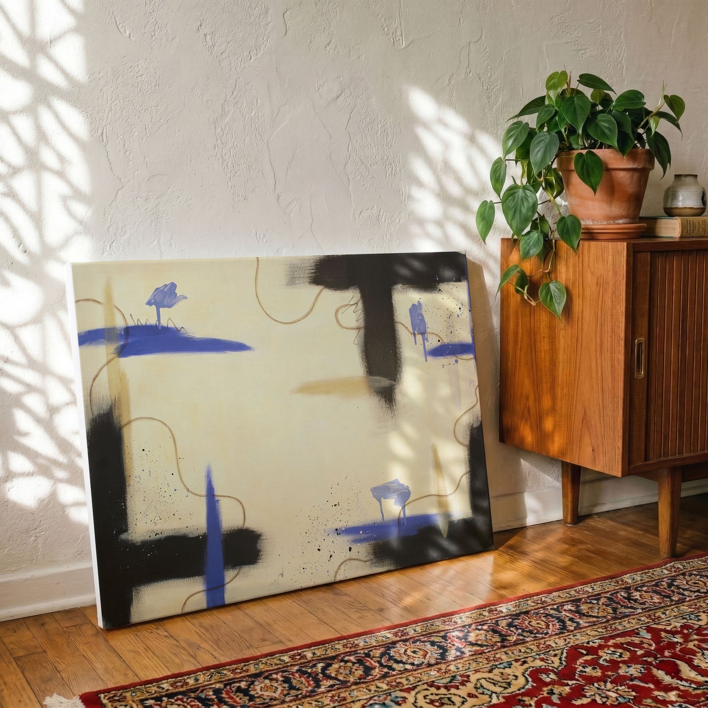 A canvas print of the painting sitting on a dark wooden floor, leaning against a textured white wall. The canvas sits in front of a red patterned rug, with a wooden armoire ont he right. On the armoire is a potted plant, a book, and grey decorative vase.