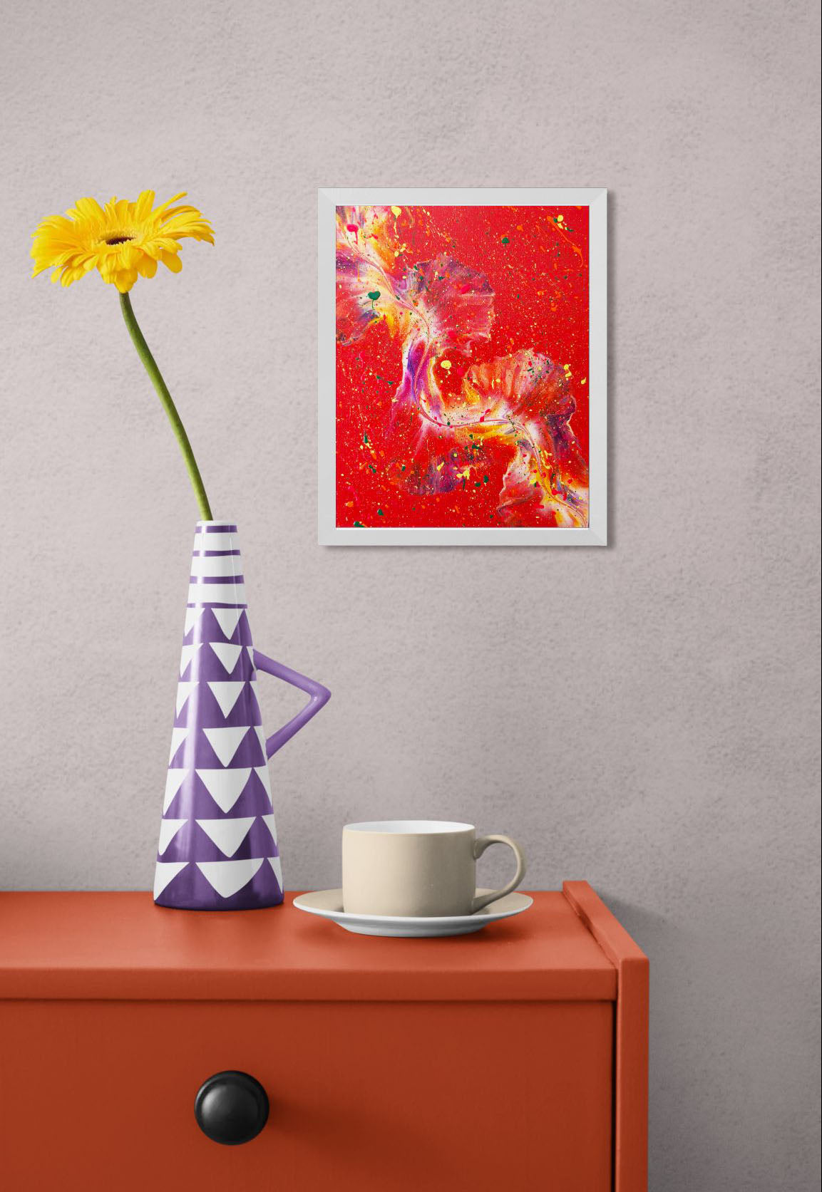 A vivid red abstract painting with bold splashes of color, framed in white, displayed above an orange side table with a beige cup and a purple geometric vase holding a yellow daisy