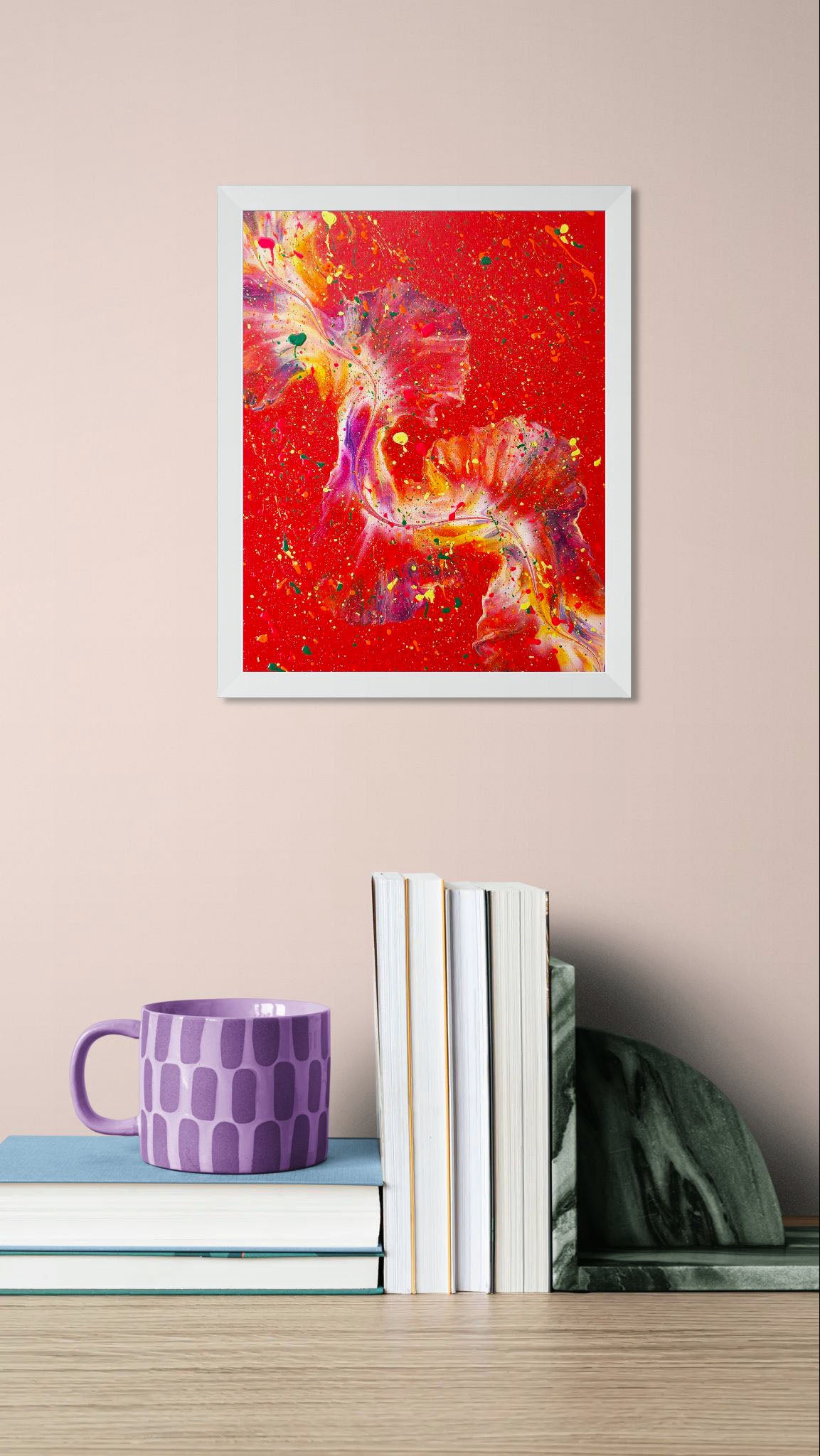 Colorful abstract painting with a fiery red background, framed in white, hanging above a stack of books and a purple mug on a wooden desk with a soft pink wall.