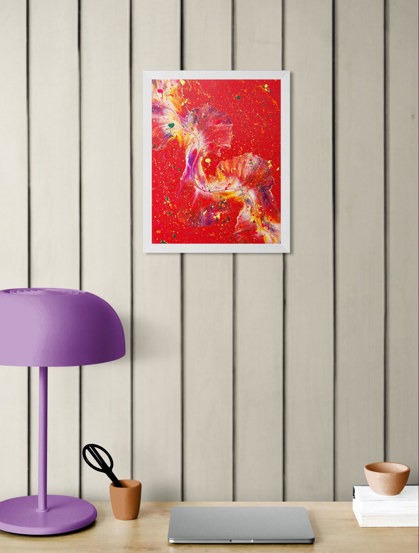 Abstract painting with bright red tones and vibrant color accents, framed in white, displayed in a modern workspace with light wooden paneling, a purple desk lamp, and a laptop.