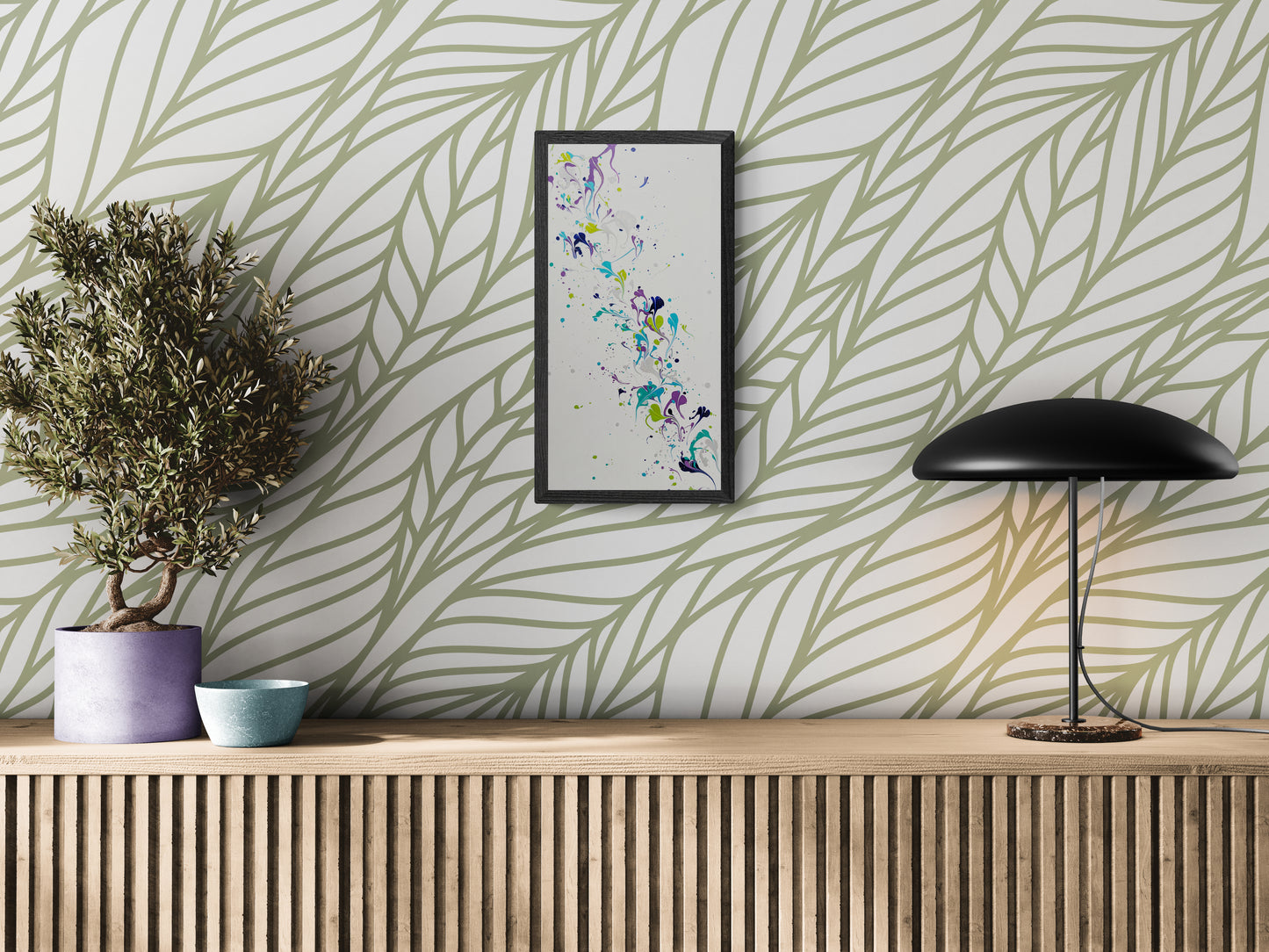A framed vertical abstract painting with flowing swirls of purple, teal, green, and silver hangs on a white wall with a large sage green leaf pattern. The artwork is displayed above a wood slatted console holding a purple planter with a small tree, a blue ceramic bowl, and a sleek black lamp. The space feels modern, airy, and rooted in natural elements.