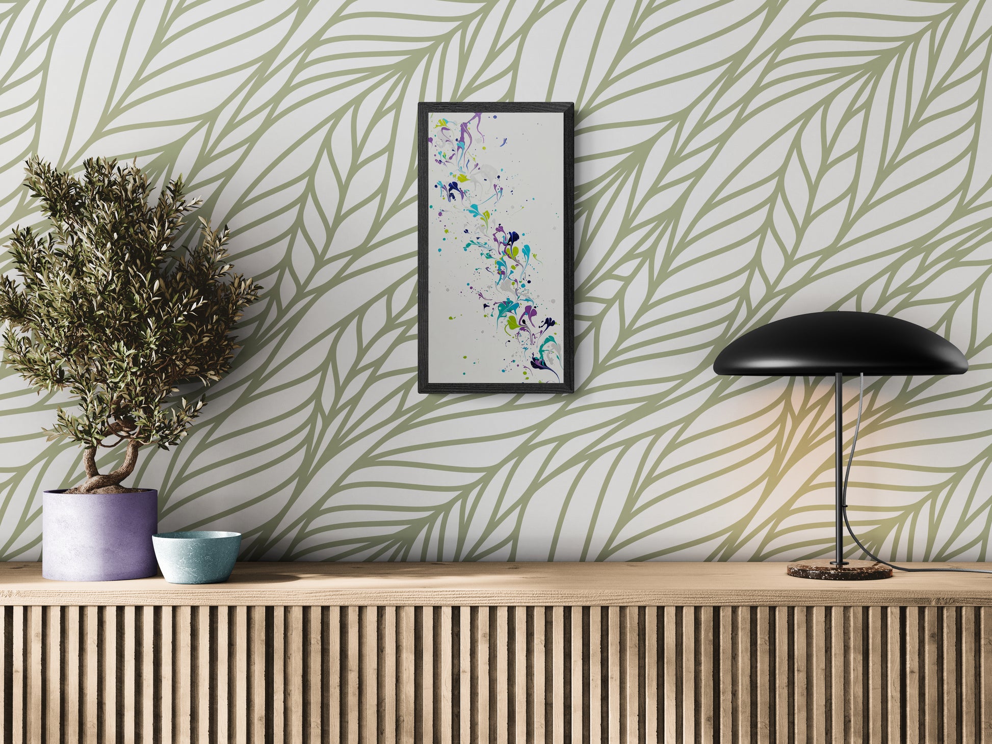 A framed vertical abstract painting with flowing swirls of purple, teal, green, and silver hangs on a white wall with a large sage green leaf pattern. The artwork is displayed above a wood slatted console holding a purple planter with a small tree, a blue ceramic bowl, and a sleek black lamp. The space feels modern, airy, and rooted in natural elements.