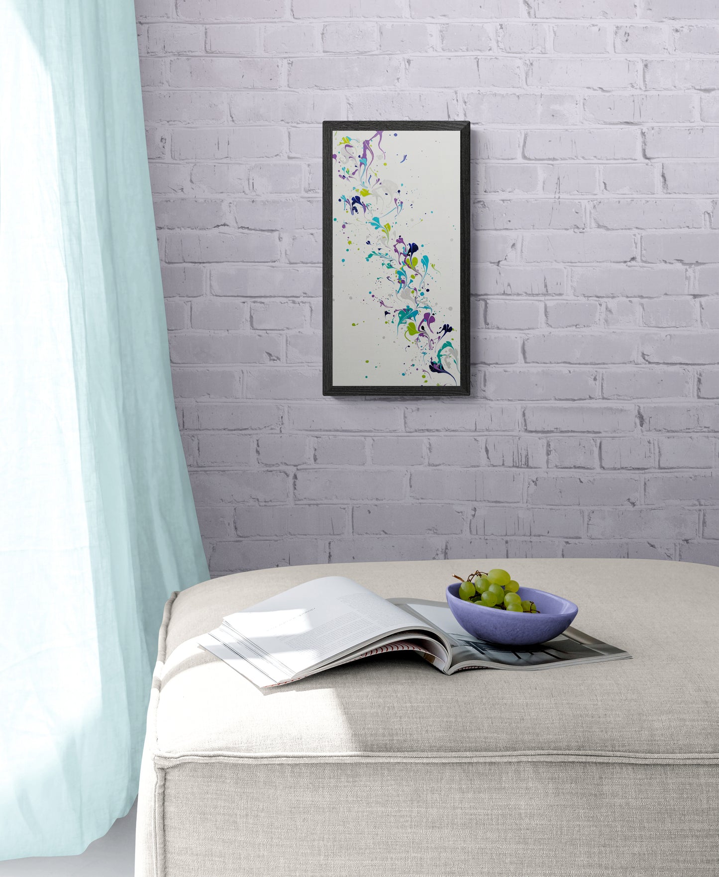 A framed abstract painting with flowing streaks of purple, teal, green, and silver hangs on a light lavender brick wall. The artwork is displayed above a cream ottoman, which holds an open magazine and a purple bowl of grapes. Soft aqua curtains frame the space, filling the room with a peaceful, gentle ambiance.