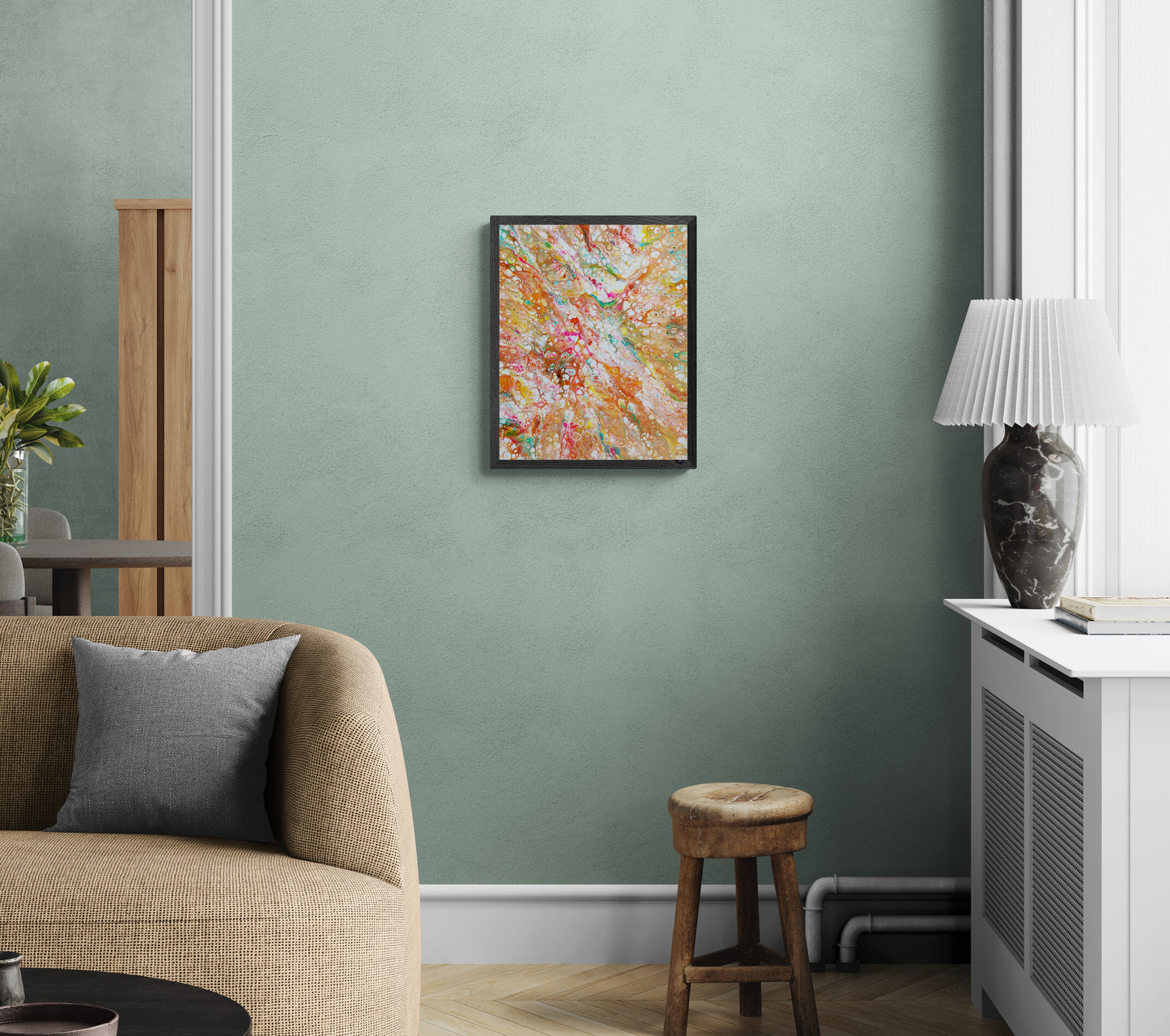 A framed abstract painting bursting with warm colors—orange, pink, teal, and gold—hangs on a soft sage green wall in a cozy living space. The room features a textured tan sofa, a gray throw pillow, a rustic wooden stool, a marble lamp on a white cabinet, and subtle natural light filtering in. The vibrant artwork adds a playful, colorful contrast to the calm, earthy palette of the room.
