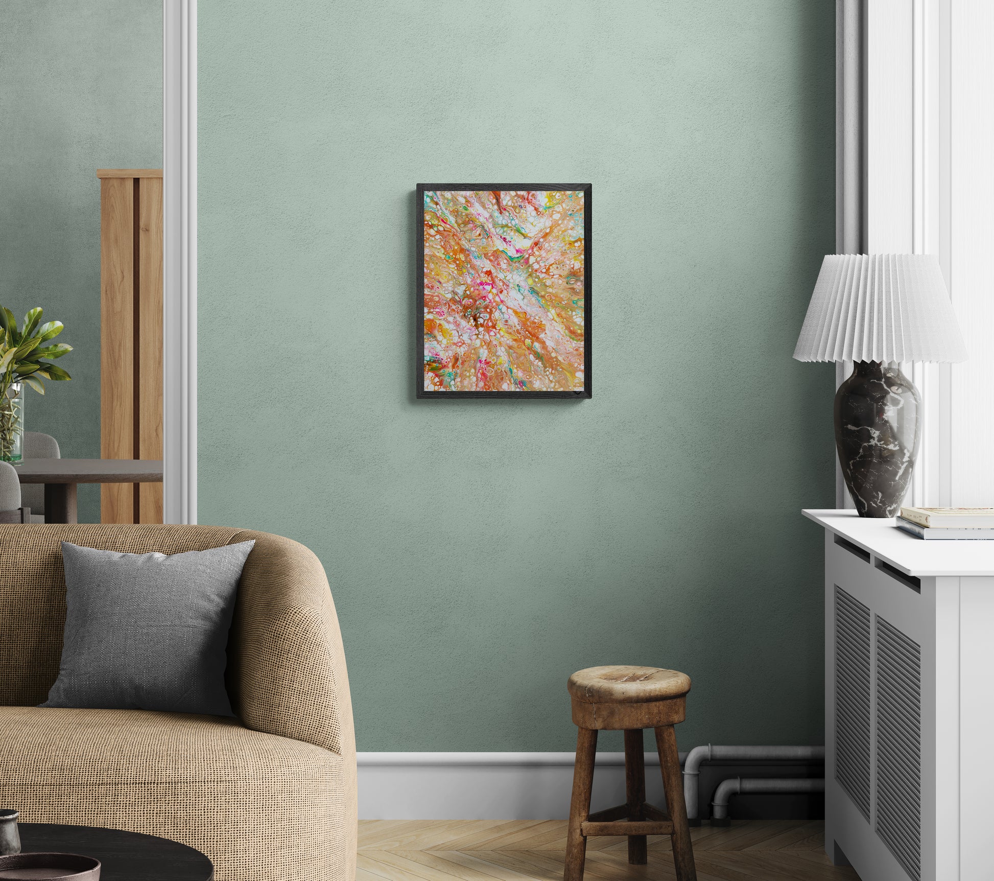 A framed abstract painting bursting with warm colors—orange, pink, teal, and gold—hangs on a soft sage green wall in a cozy living space. The room features a textured tan sofa, a gray throw pillow, a rustic wooden stool, a marble lamp on a white cabinet, and subtle natural light filtering in. The vibrant artwork adds a playful, colorful contrast to the calm, earthy palette of the room.