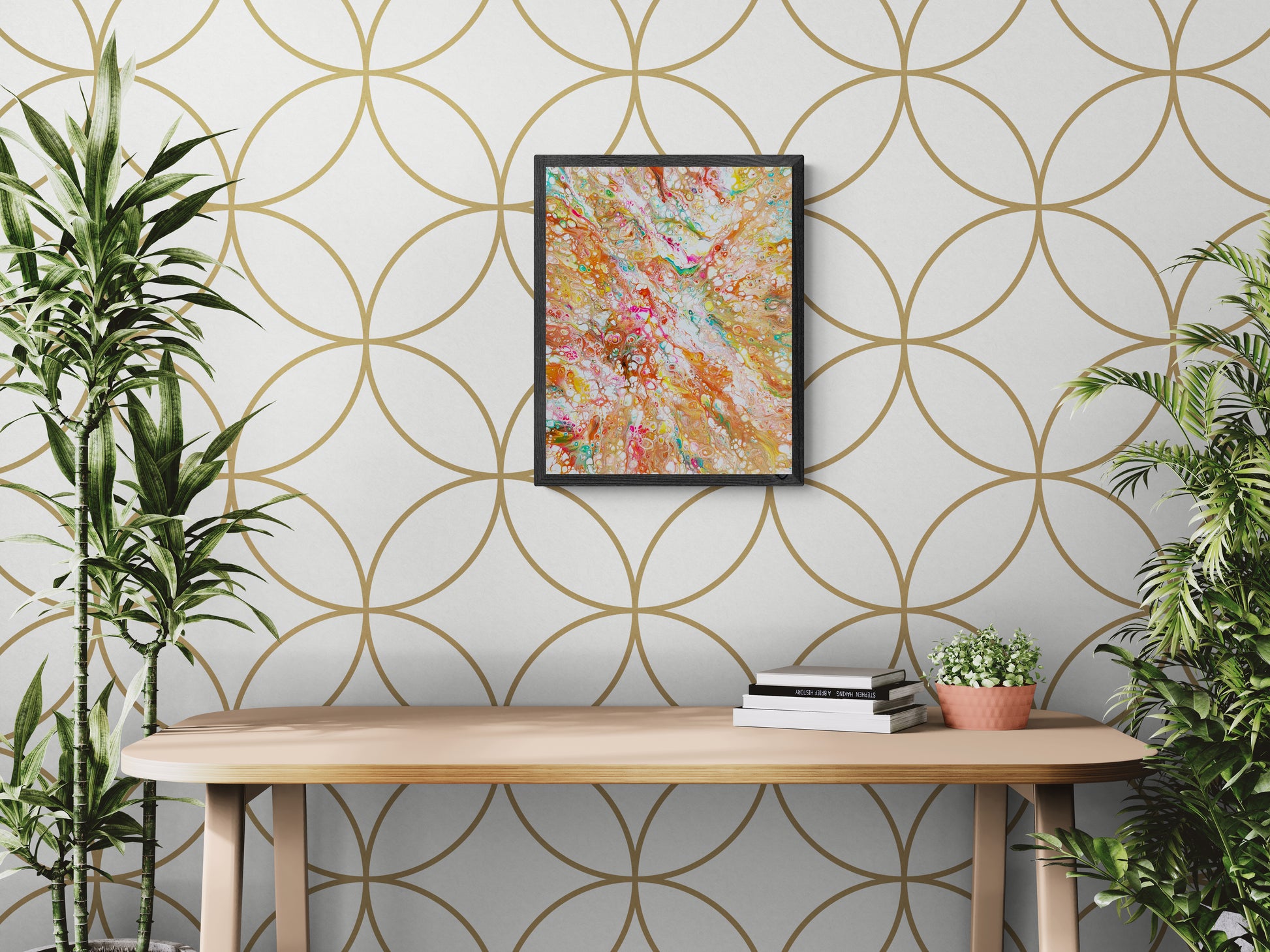 A colorful abstract painting featuring swirling hues of orange, pink, teal, and white hangs on a white wall with a large gold geometric circle pattern. Below it is a light wood desk with stacked books and a small potted plant. Lush green plants frame the scene on both sides, bringing a vibrant, natural energy to the space.