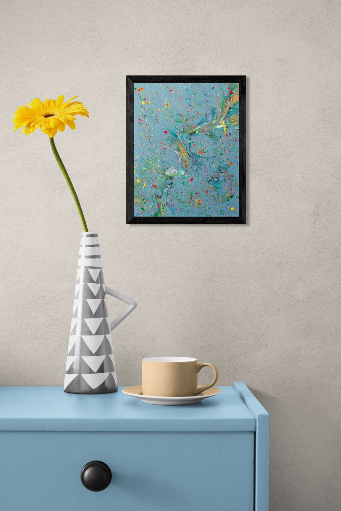 A framed abstract fluid art painting with a blue-green background and colorful splatter details, displayed above a light blue dresser. A geometric-patterned gray and white vase with a yellow flower and a beige coffee cup are placed on the dresser.