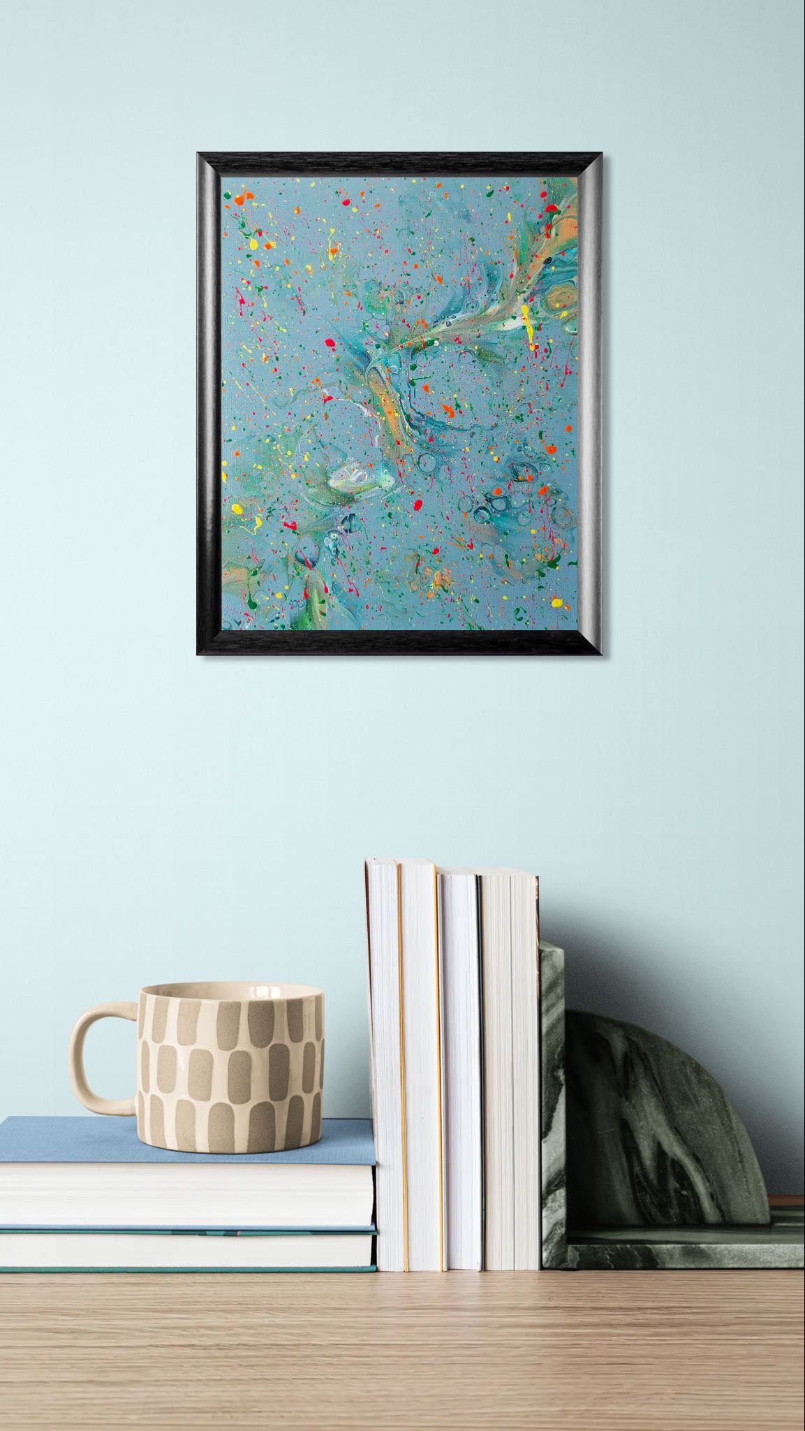 A framed square abstract painting with swirling blue-green tones and vibrant paint splatters in yellow, orange, pink, and green, shown hanging above a stack of books and a beige mug with a geometric pattern. The background wall is pale blue.
