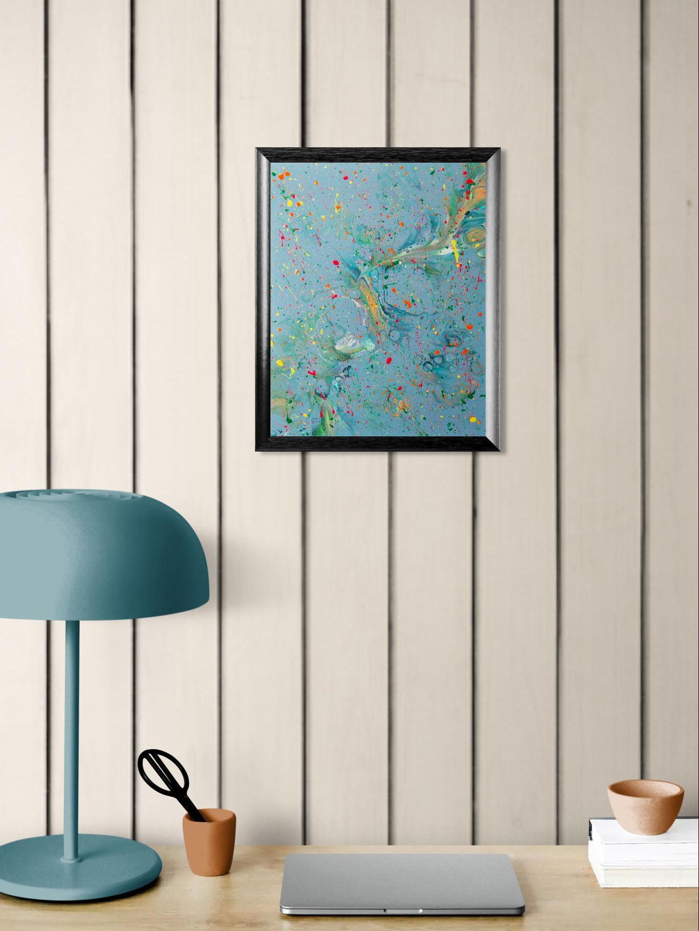 Alt Text: A framed abstract fluid painting with a soft teal-blue background and bursts of colorful splatter in red, yellow, pink, and green. The artwork hangs above a wooden desk setup that includes a teal lamp, a terracotta cup with scissors, and a closed laptop.