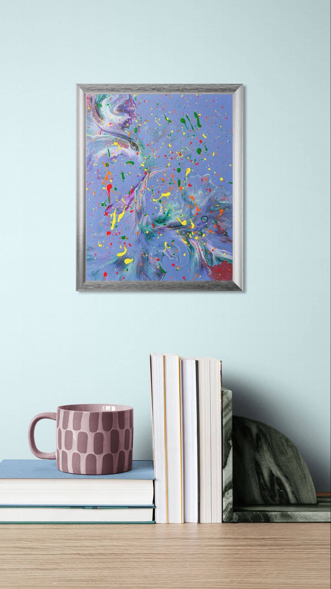 Mockup of the painting displayed on a pale blue wall above a stack of books with a mauve patterned mug. The vibrant artwork stands out with its bold splatters and swirling color movement