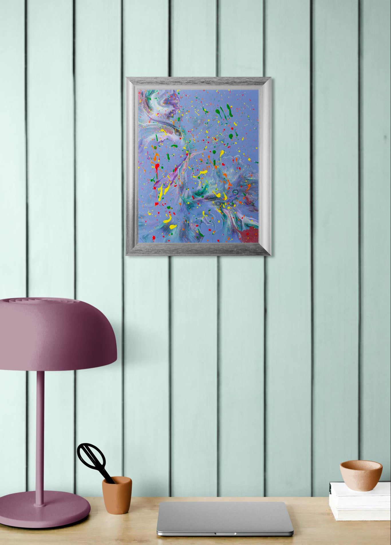 Mockup of the painting hung on a wood-paneled wall above a desk setup with a mauve lamp, terracotta cup, and closed laptop. The colorful painting brings a lively, artistic energy to the workspace