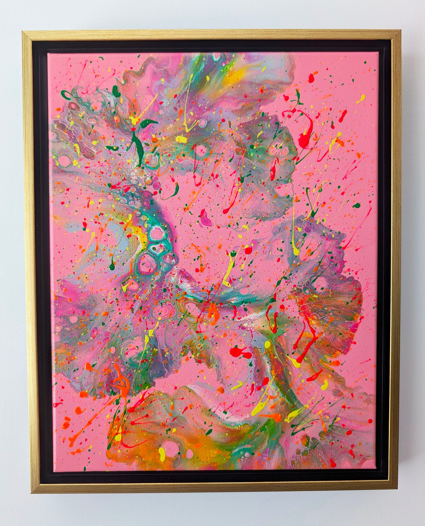 Framed acrylic fluid art painting with a vivid pink background. Flowing marbled textures in green, orange, yellow, and purple sweep across the canvas, overlaid with energetic neon splatters in red, yellow, and green