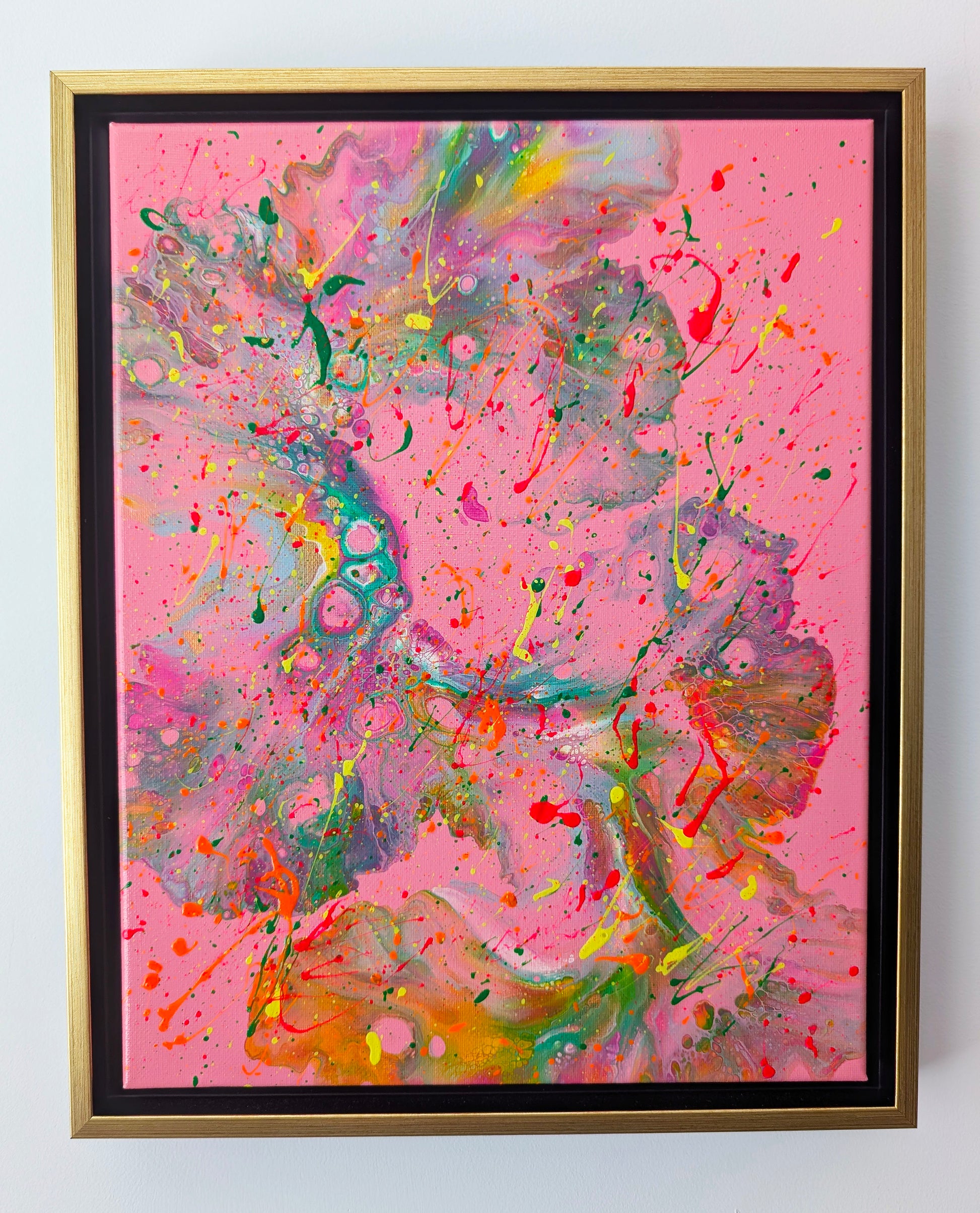 Framed acrylic fluid art painting with a vivid pink background. Flowing marbled textures in green, orange, yellow, and purple sweep across the canvas, overlaid with energetic neon splatters in red, yellow, and green