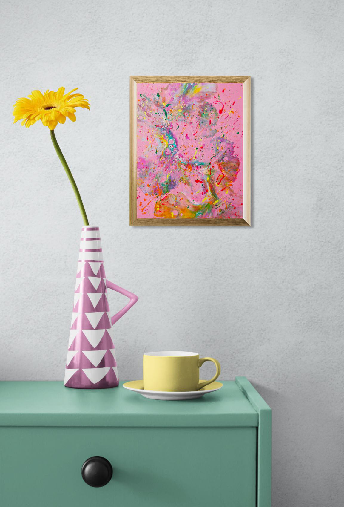 Mockup of the painting hanging on a light gray wall above a mint green cabinet. A pink and white geometric vase with a yellow flower and a yellow cup and saucer sit on the cabinet, complementing the playful tones of the artwork.
