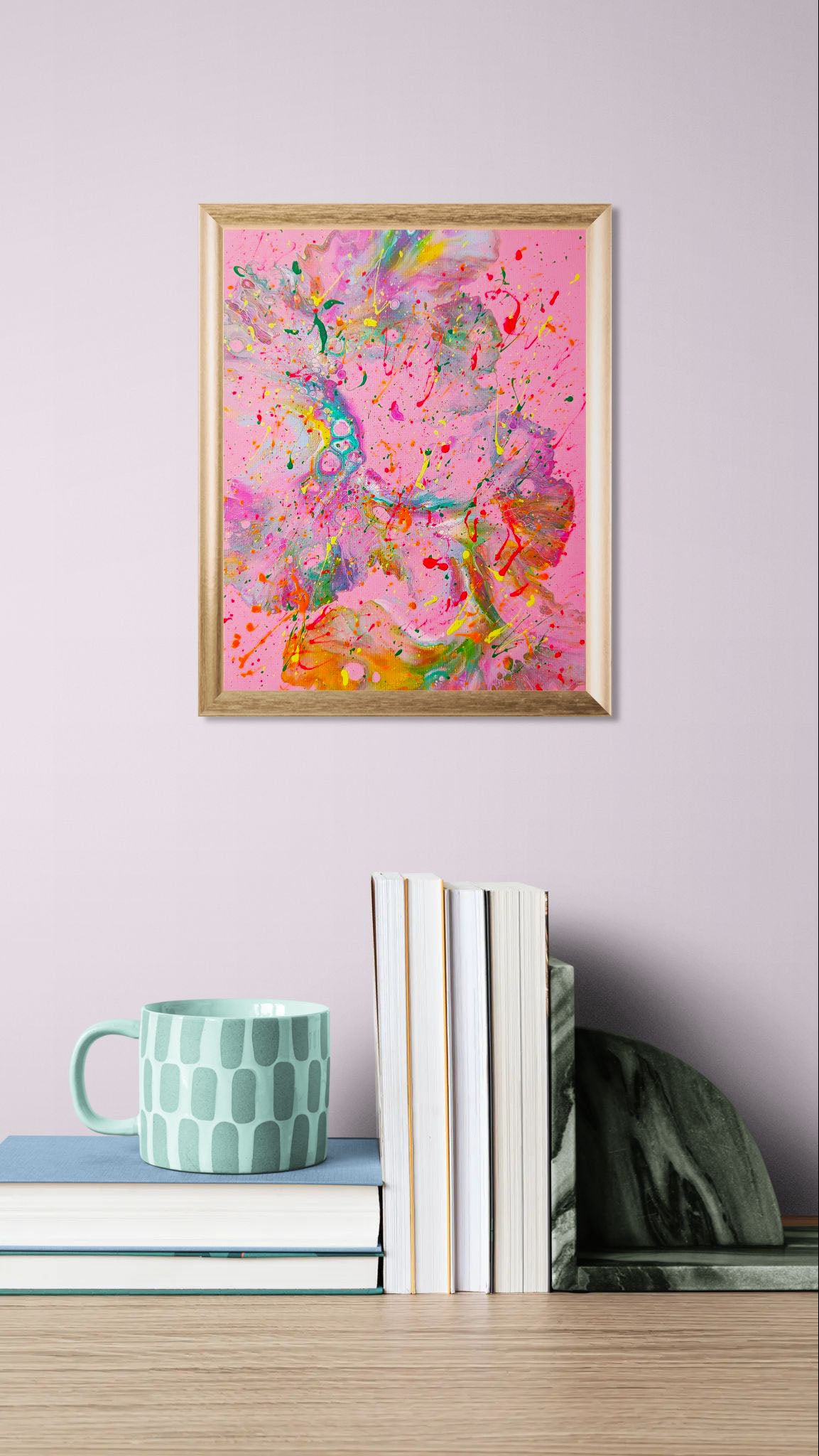 Mockup of the painting on a soft lavender wall above a stack of books with a pale mint patterned mug. The artwork adds a vibrant burst of pinks, oranges, and greens to the calm, minimalist space.