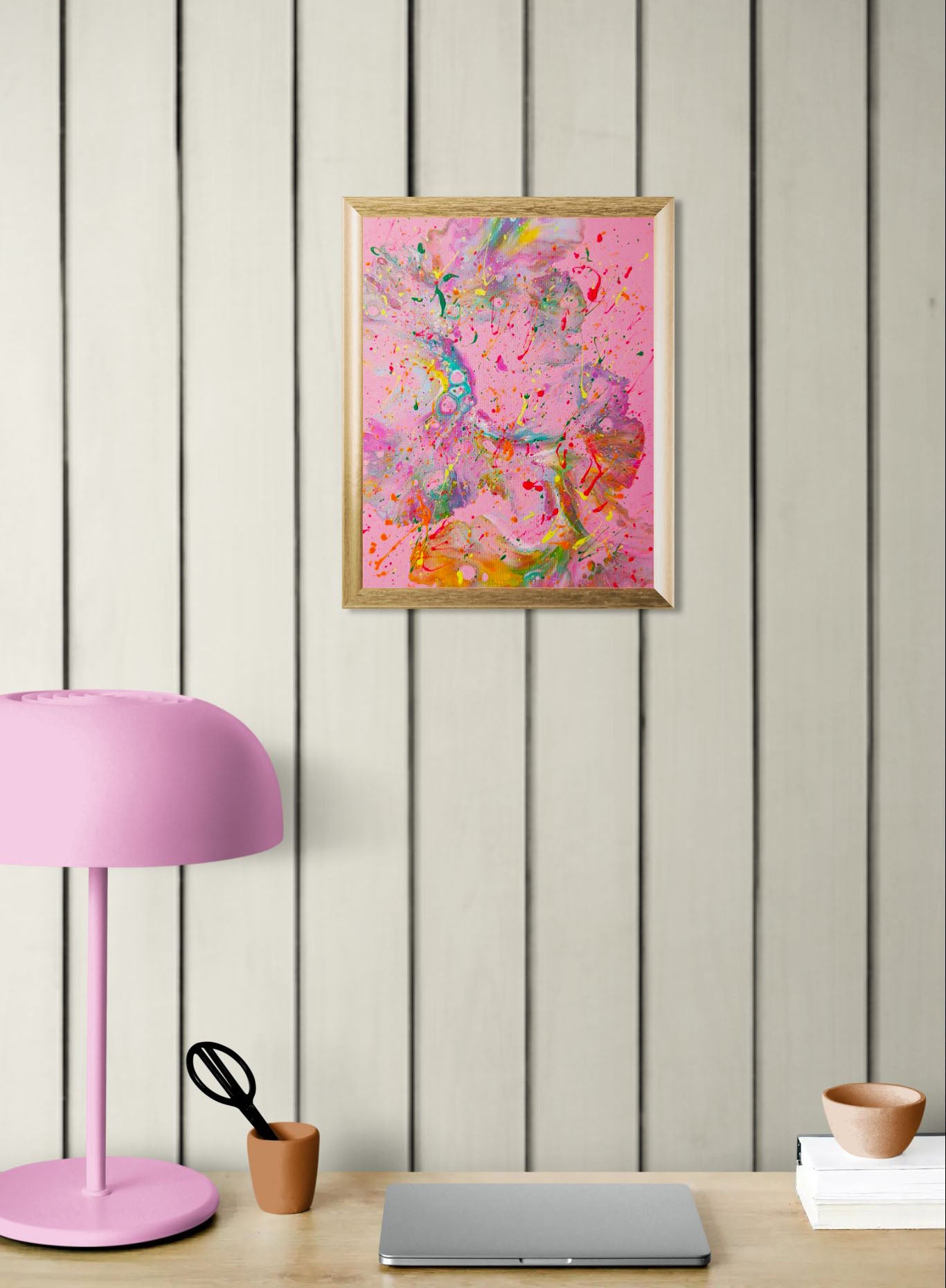 Mockup of the painting hung on a wood-paneled wall above a desk with a pink lamp, terracotta cup, and a closed laptop. The bold and colorful painting adds an expressive, energetic focal point to the workspace