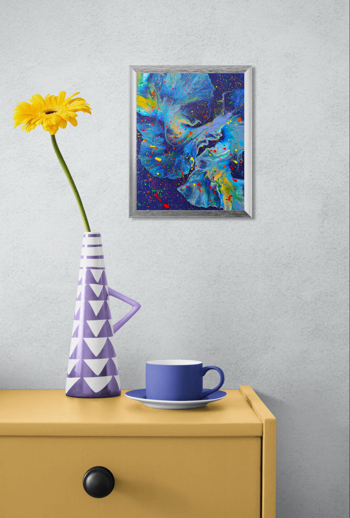 Framed deep blue and dark purple fluid painting with vibrant splashes of red, yellow, green, and orange, displayed above a mustard-colored dresser.