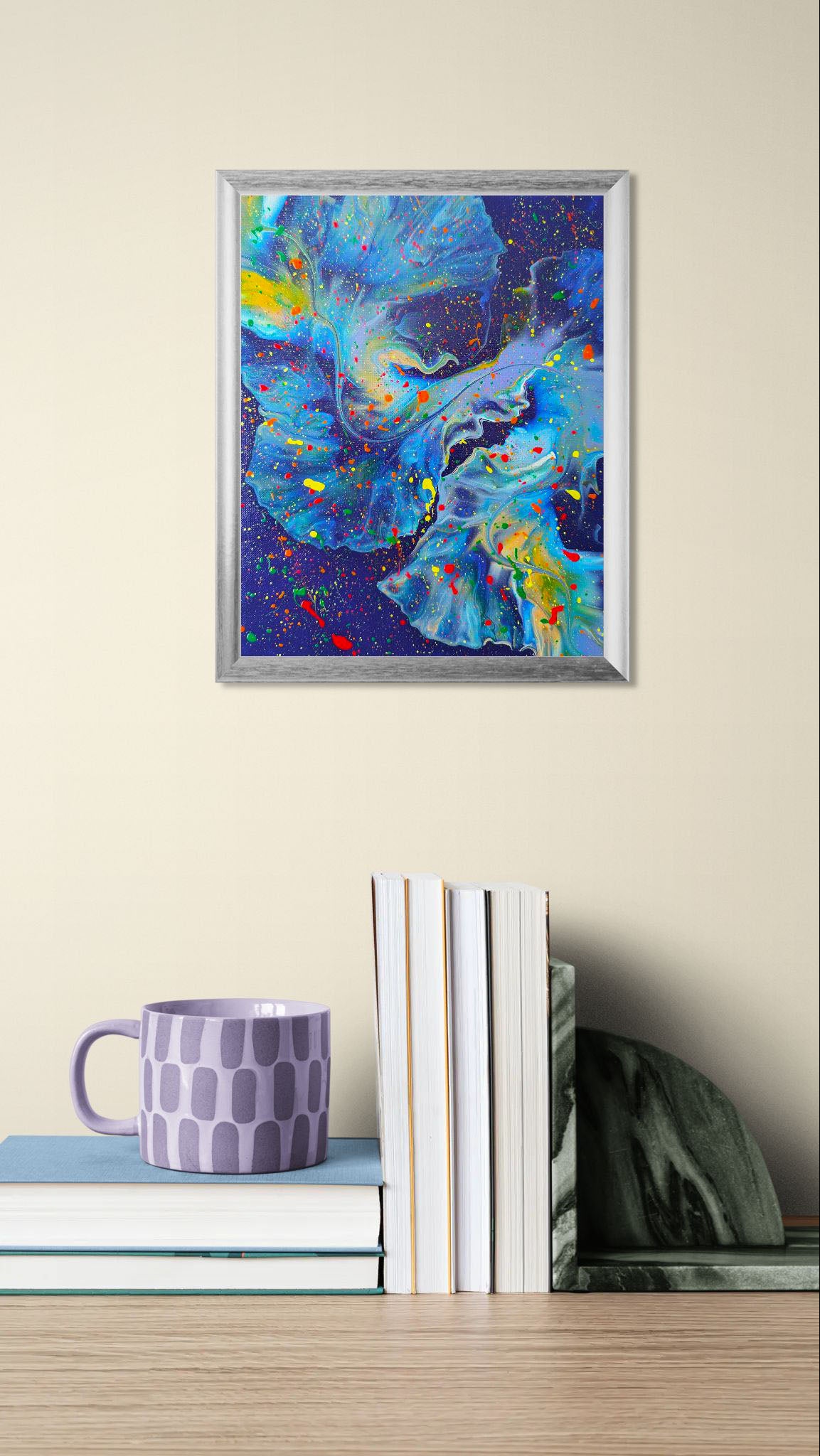 Cosmic blue abstract fluid painting with swirling textures and bold splatters of neon red, yellow, green, and orange, shown above stacked books with a lavender mug.