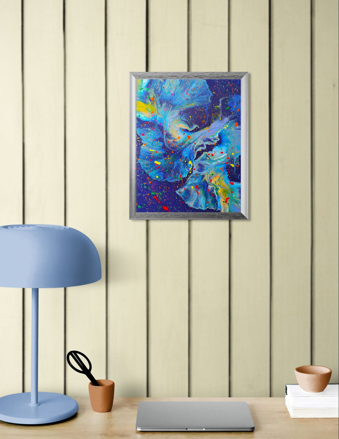 Framed deep blue and purple abstract painting on a neutral paneled wall, styled with a pale blue lamp, laptop, and minimalist desk accessories.