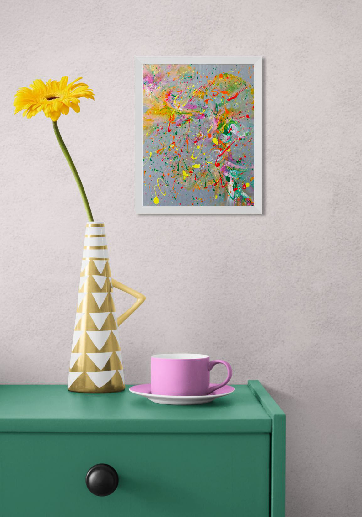Framed abstract fluid painting with bright neon splatters over a gray background, displayed above a green side table with a geometric yellow vase and a pink teacup.