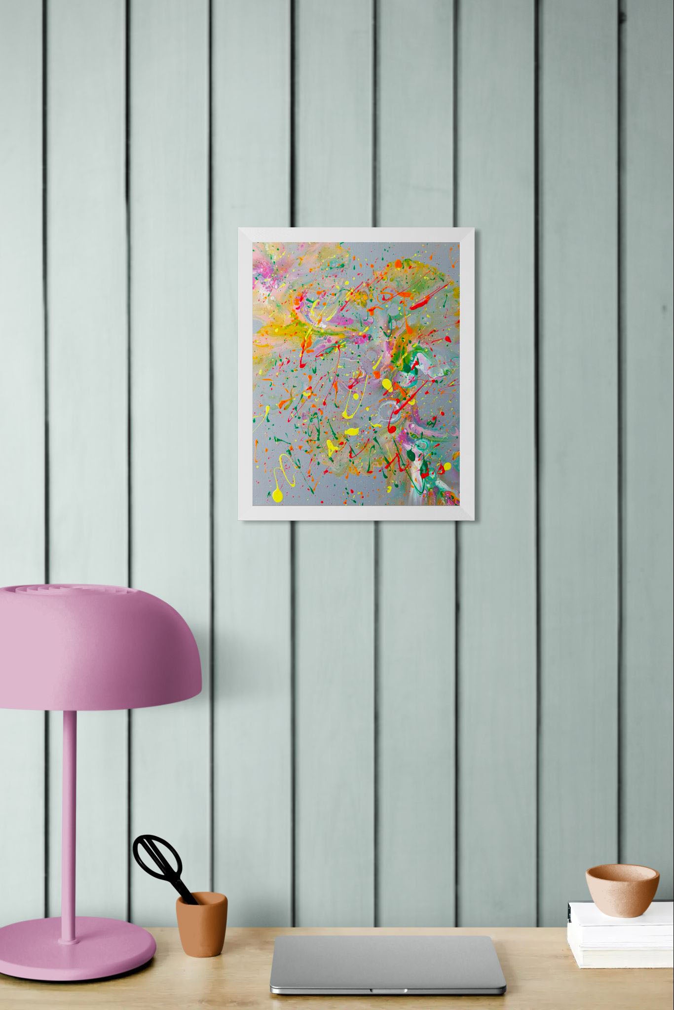 Abstract fluid painting in a white frame with vibrant splashes of color on a gray background, hanging above a desk setup with a pink lamp, scissors, and a laptop.