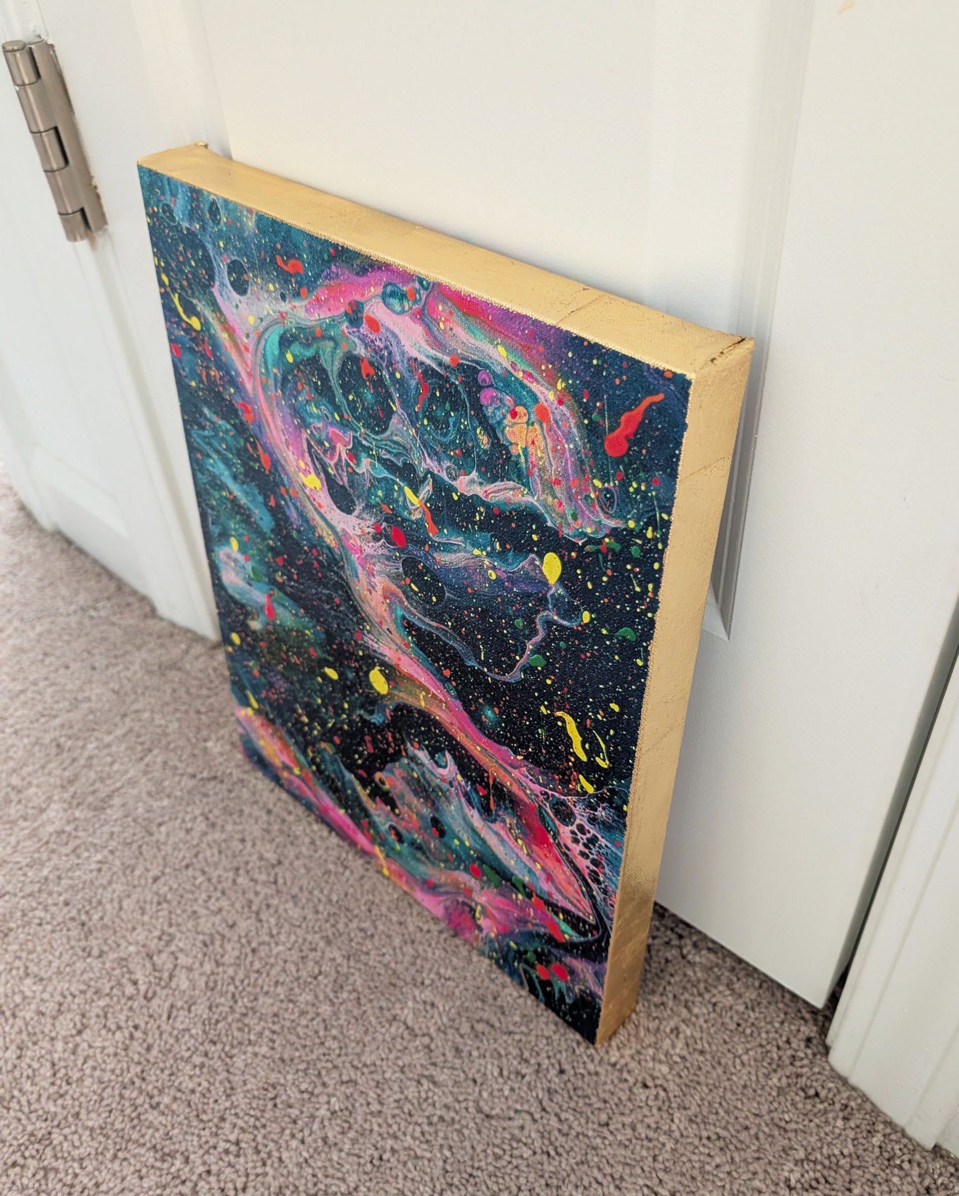 Close angled view of the canvas leaning against a door, showing the metallic gold leaf edge next to the bold teal, pink, and yellow fluid patterns.