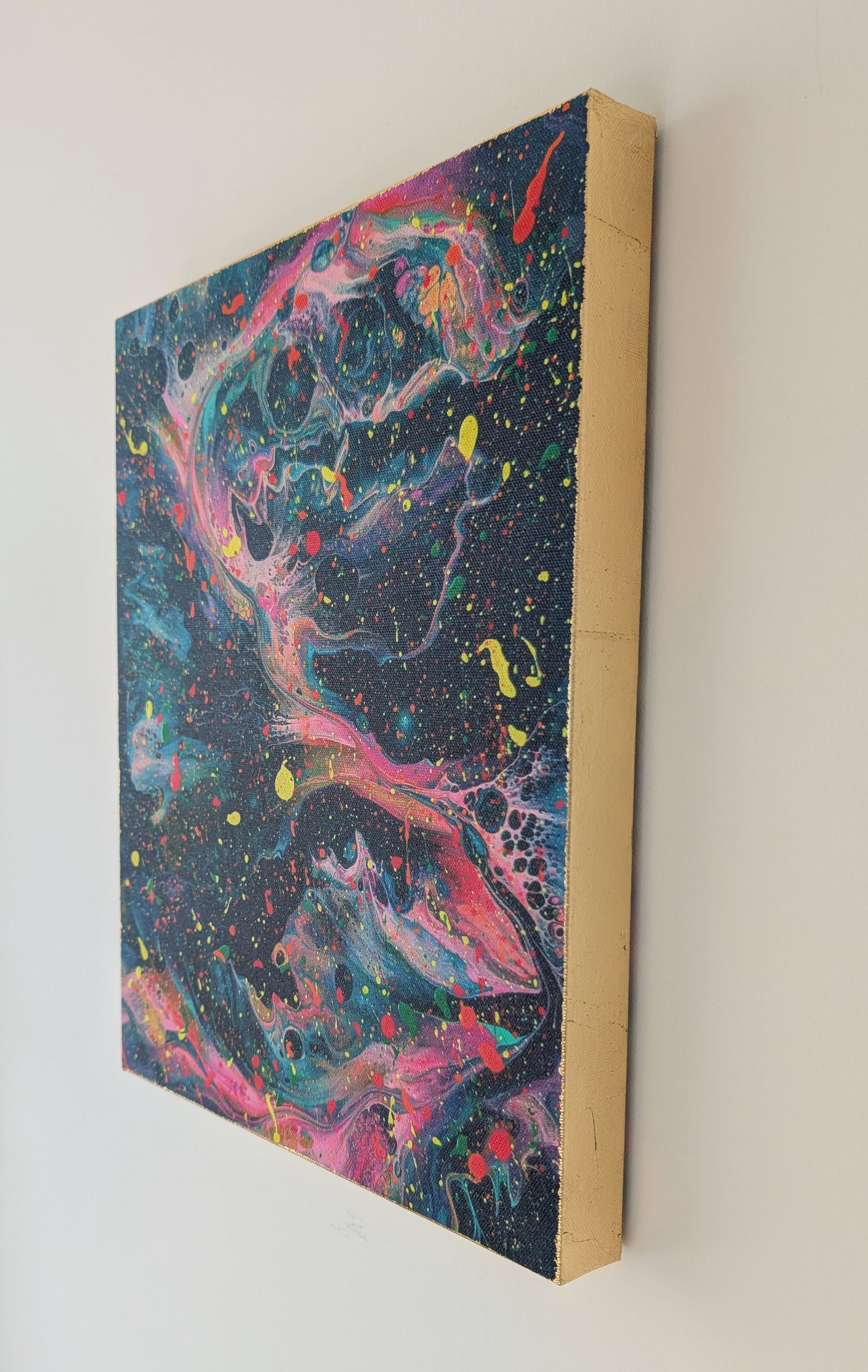 Angled close-up of the canvas on a wall, emphasizing the hand-applied gold leaf edge and the colorful layered fluid patterns on the deep background.