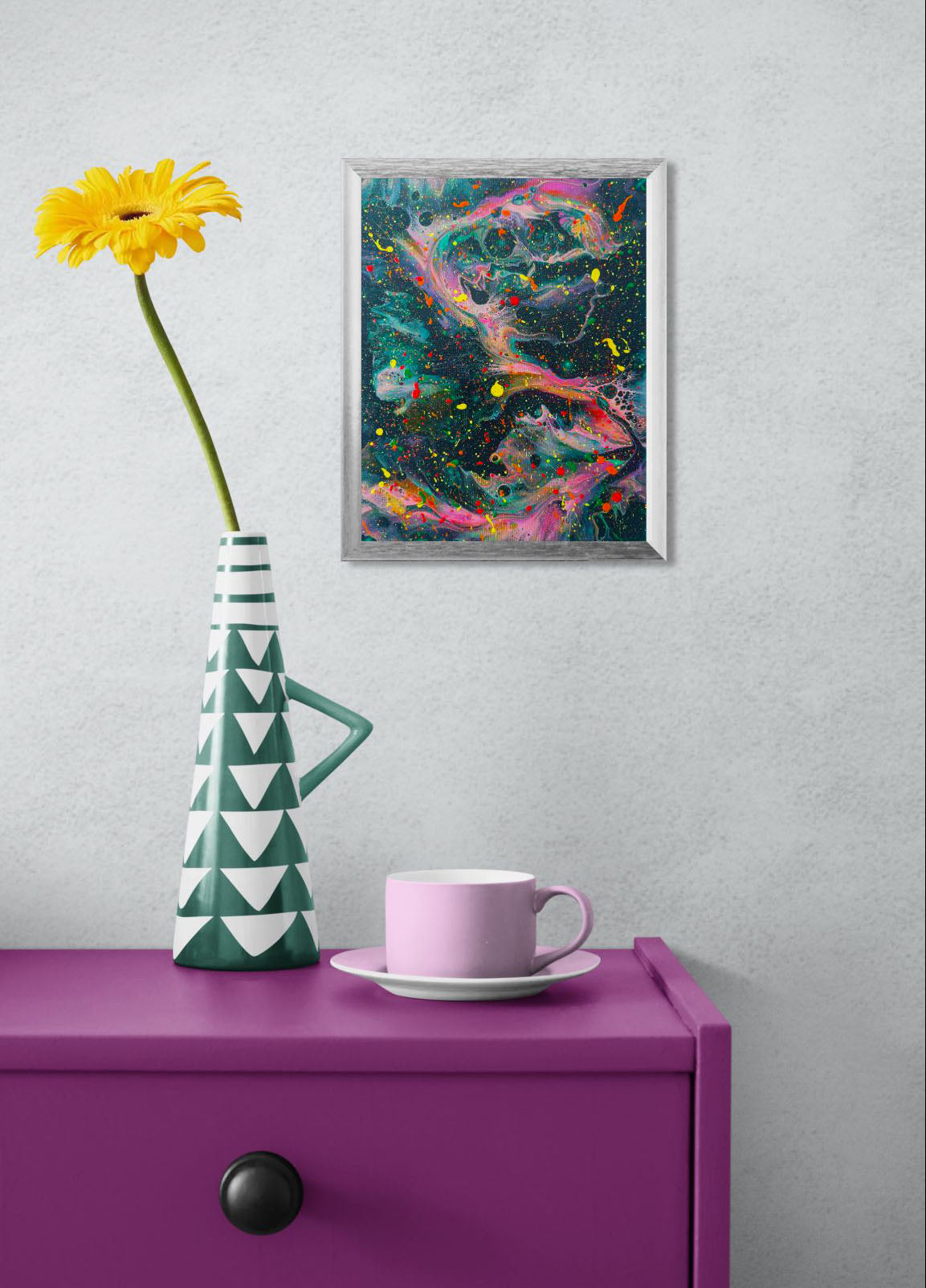 Framed abstract fluid painting with a dark green background, bright neon pink swirls, and vivid splatters of yellow, red, and green. The artwork hangs on a light gray wall above a bold purple side table with a geometric green and white vase and a soft pink teacup.
