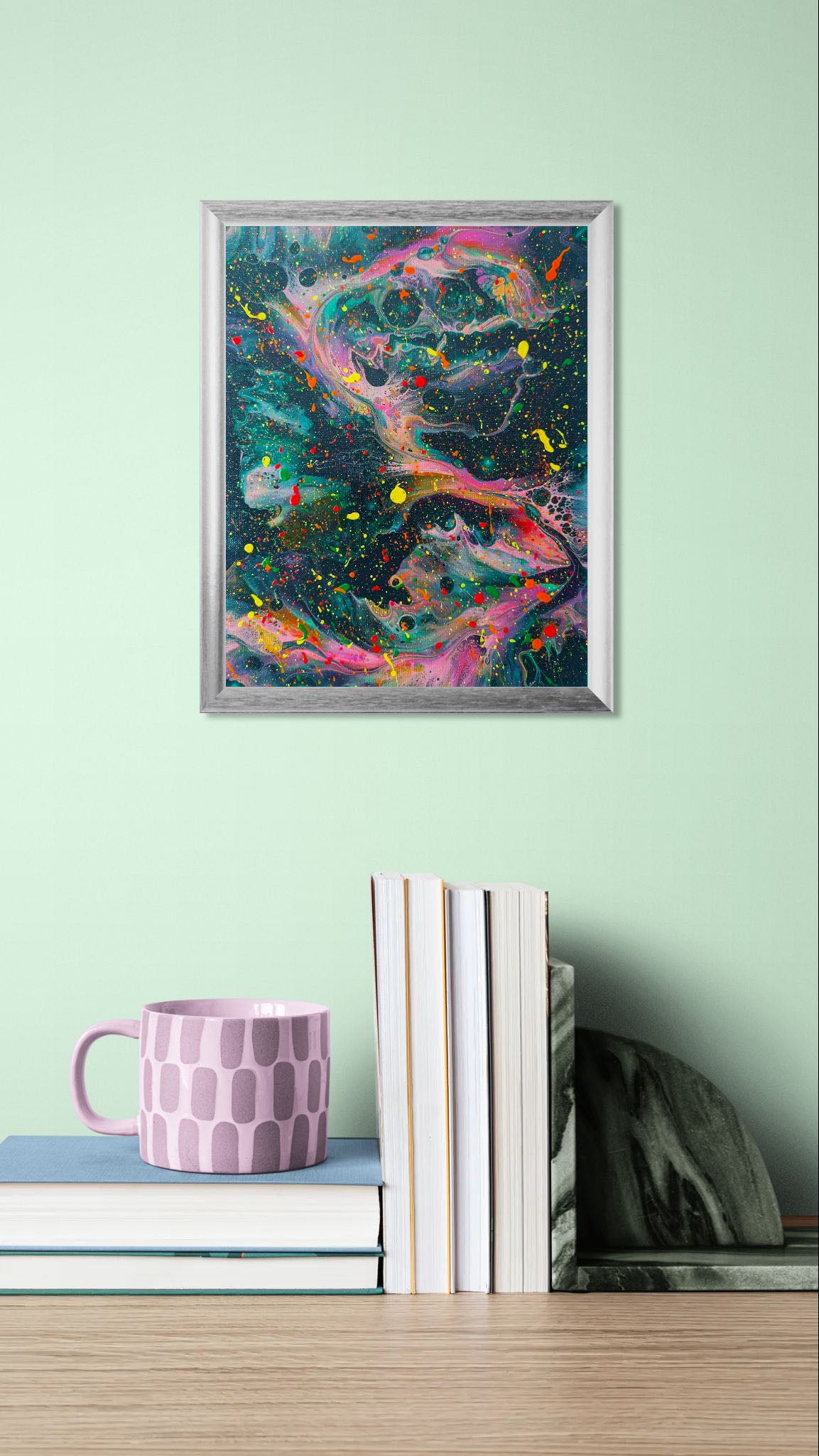 Framed abstract painting featuring vibrant neon pink and orange swirls over a deep green base, with scattered splashes of yellow, red, and green. Displayed on a mint green wall above a wooden desk with stacked books and a pink mug.