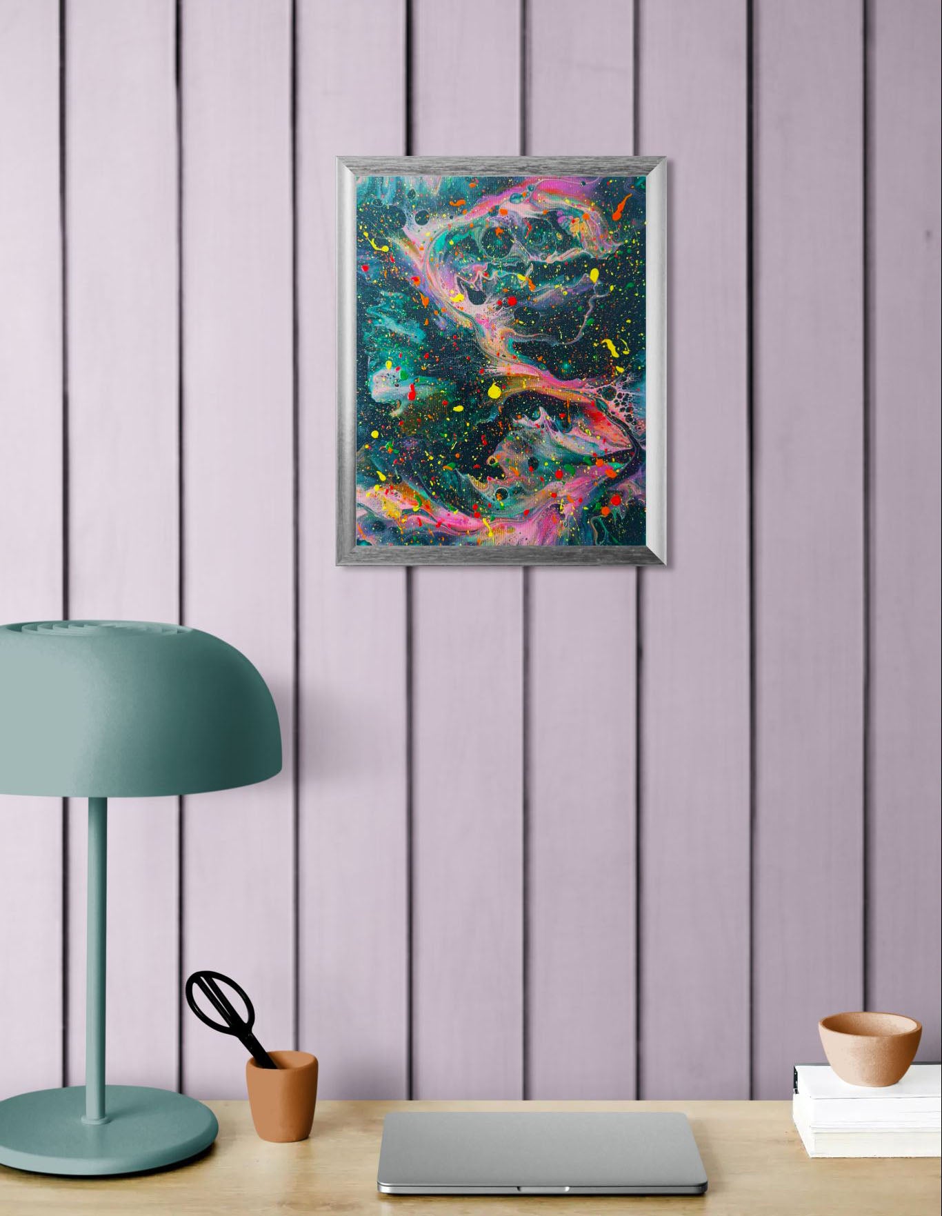 Framed abstract fluid art piece with a dark green background, dynamic neon pink and orange streaks, and high-contrast paint splatters. Shown against a lavender wooden plank wall above a desk setup with a muted teal lamp and small bowls