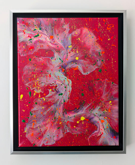 A close-up of a vibrant fluid art painting featuring a deep red background with flowing forms in lavender, purple, and teal. Bright splatters of yellow, orange, neon pink, and green add energetic contrast. The painting is framed in a silver and black floating frame.