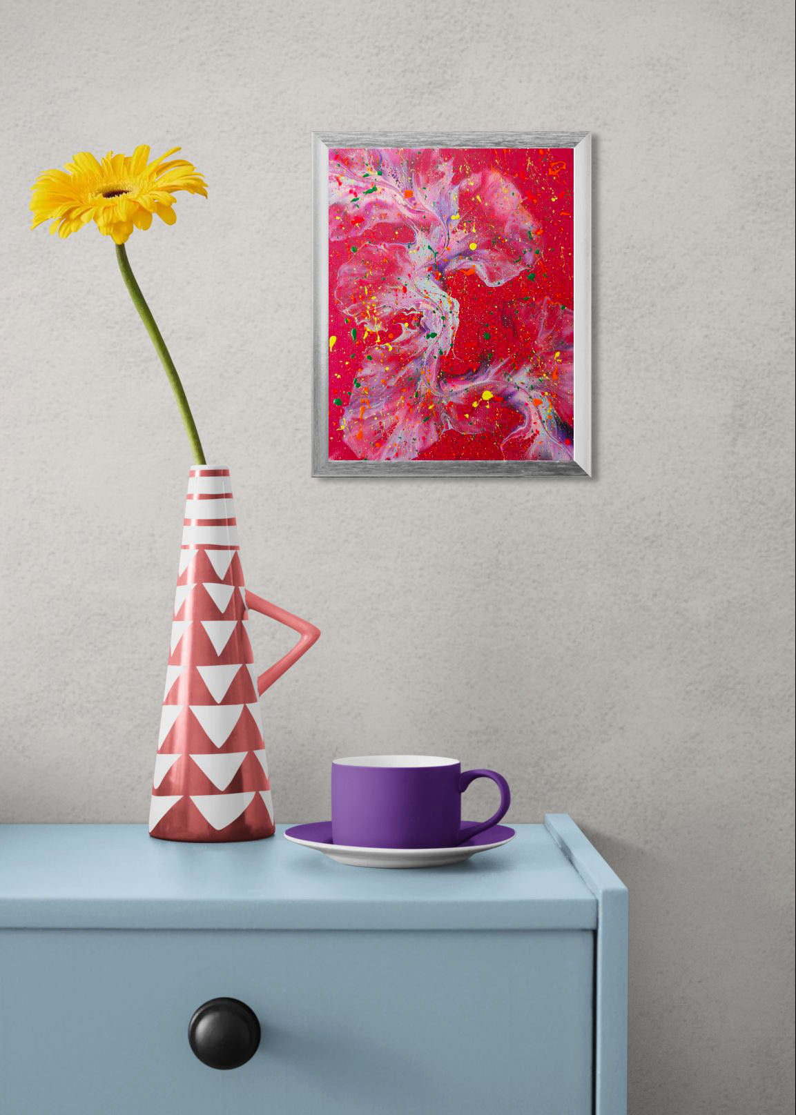  framed abstract fluid art painting with a deep red background and swirling accents of purple, blue, and green, splattered with neon yellow, orange, and pink. The artwork hangs above a light blue cabinet with a geometric pink vase holding a yellow daisy and a bold purple teacup