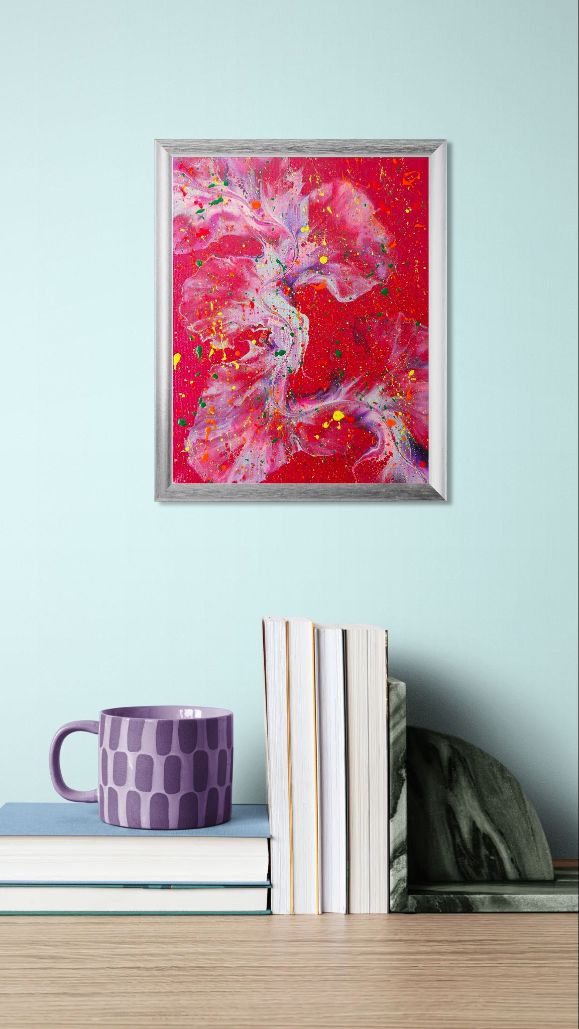 A colorful fluid art painting with a dramatic red background, layered with swirling forms of purple and blue, accented by bright splatters of yellow, orange, and green. The painting is framed and displayed on a mint green wall above a stack of books and a lavender patterned mug.