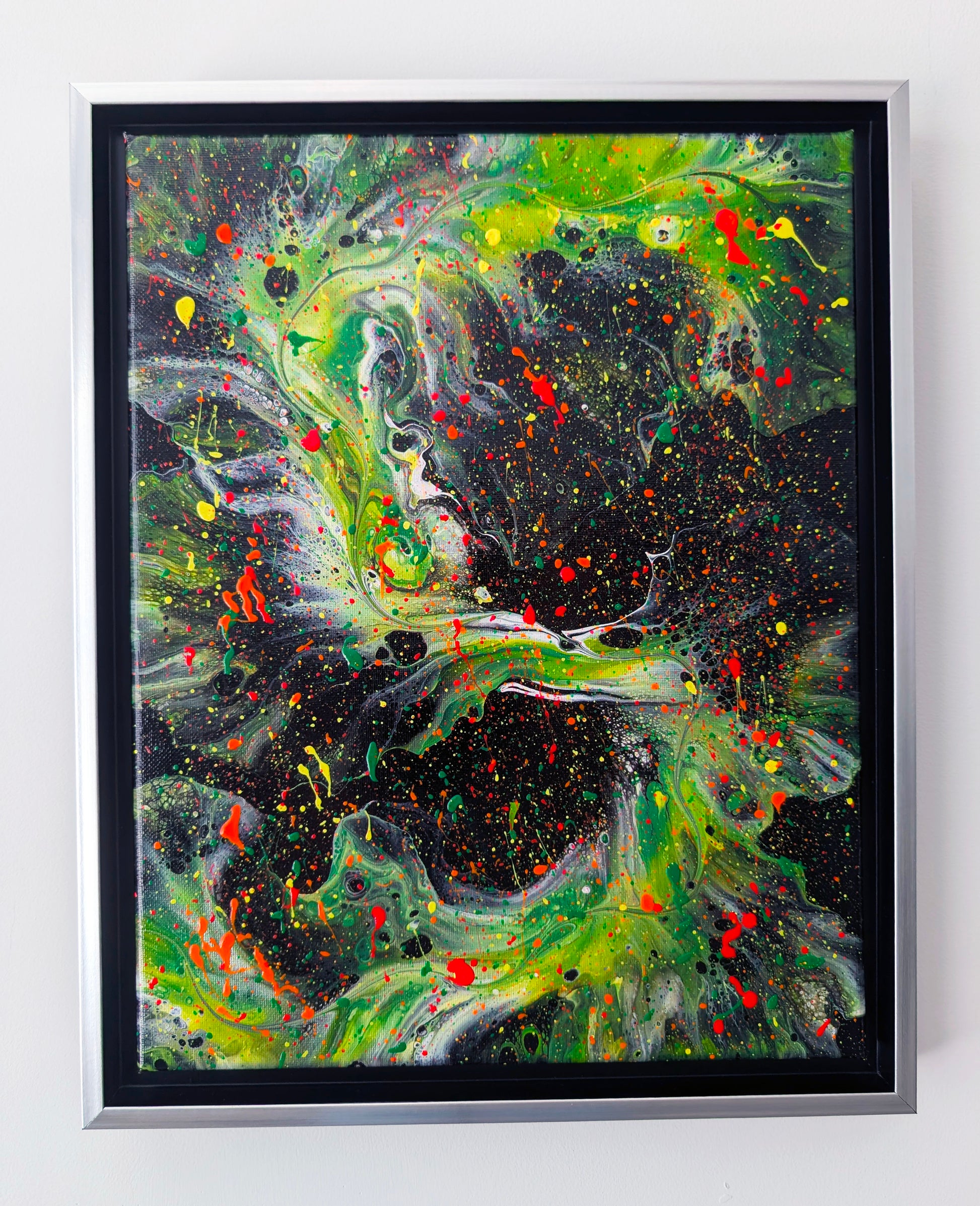 Abstract acrylic fluid painting with vibrant green, yellow, and red splatters swirling over a deep black background. The texture features dynamic movement and speckled layers, framed in silver with a black inset.