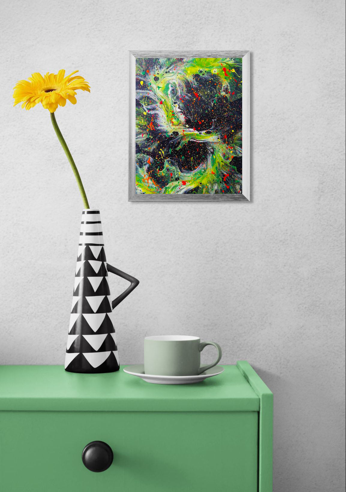 Framed abstract fluid art painting hanging above a green table with a black and white geometric vase holding a yellow flower and a pale green teacup. The painting has a black background with neon green, red, and yellow splatters and swirling patterns.