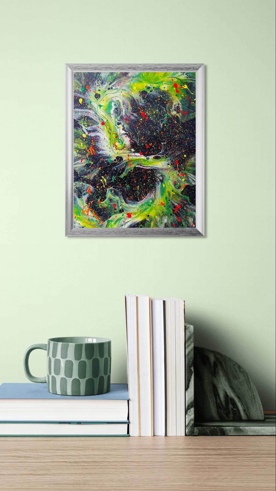  Framed fluid art painting with a cosmic, electric green and black color palette, accented by vivid red and yellow splatters. Displayed on a desk with stacked books, a patterned green mug, and a stone bookend.