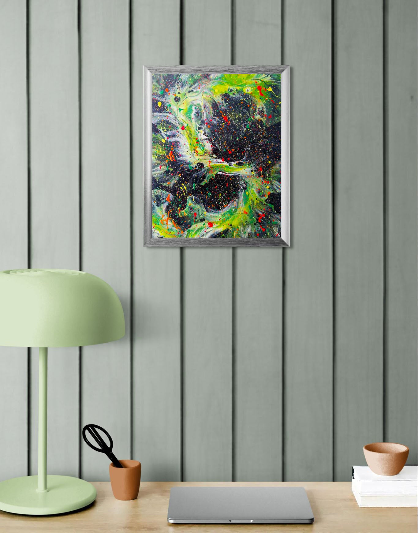 Abstract painting featuring swirling neon green forms on a black background, with bold splashes of red, yellow, and orange. Shown in a silver frame above a mint green desk with a pale green lamp and simple office accessories.