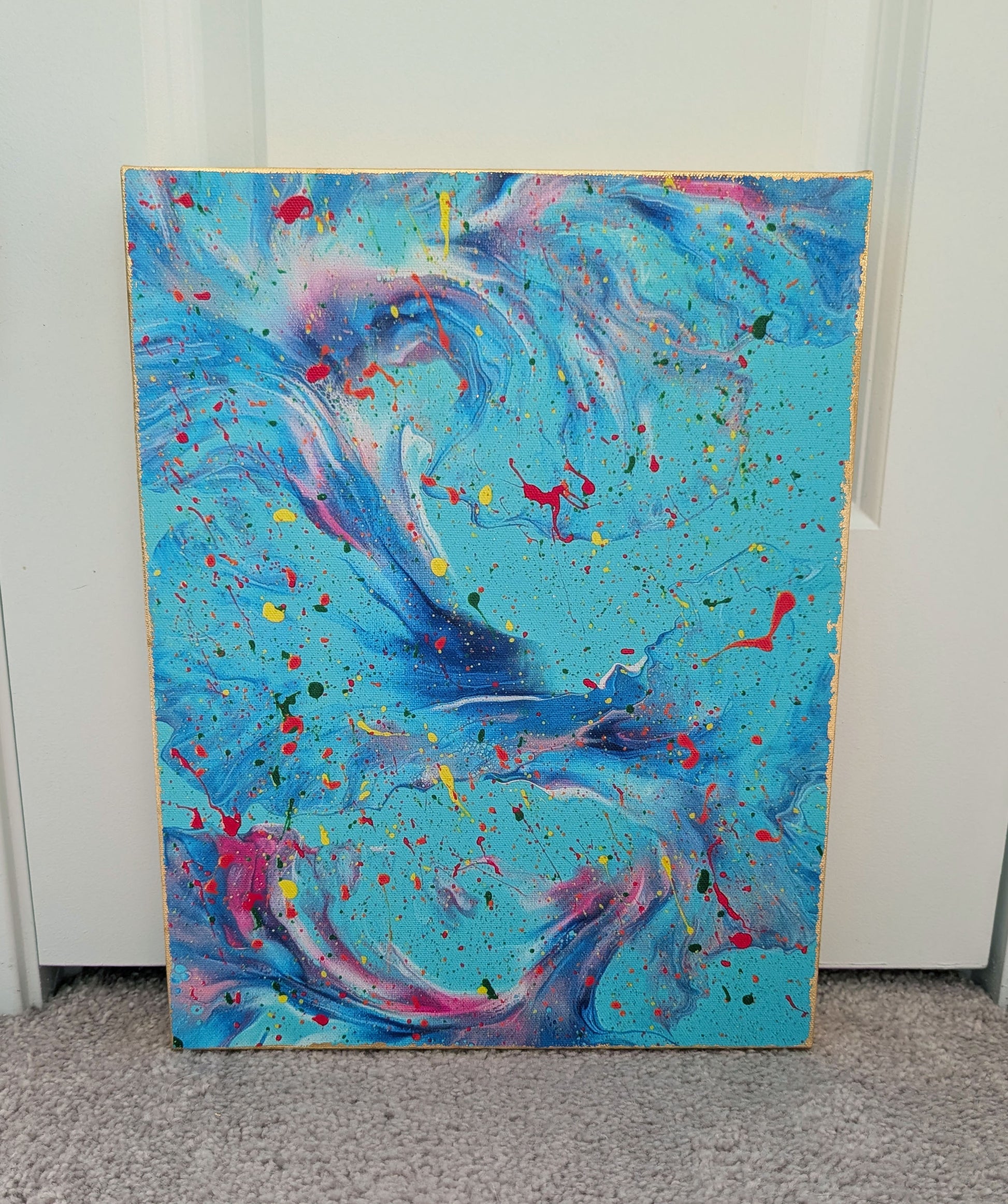 Front view of an abstract blue fluid art canvas with swirling teal, purple, pink, and blue movement, accented by colorful paint splatters. The canvas edges are finished with gold leaf.