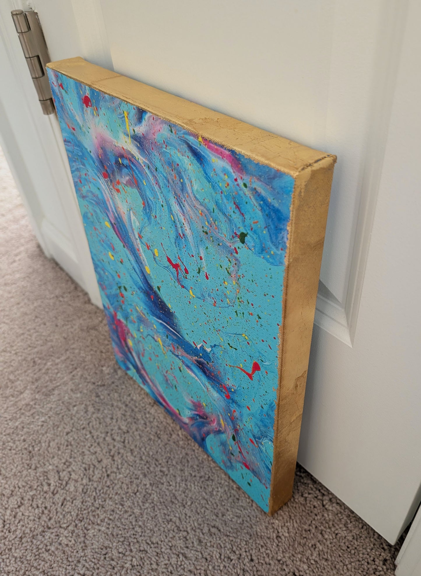 Close angled view of the blue abstract canvas leaning against a white wall, showing the gold leaf edge and swirling pink, purple, and blue fluid patterns.