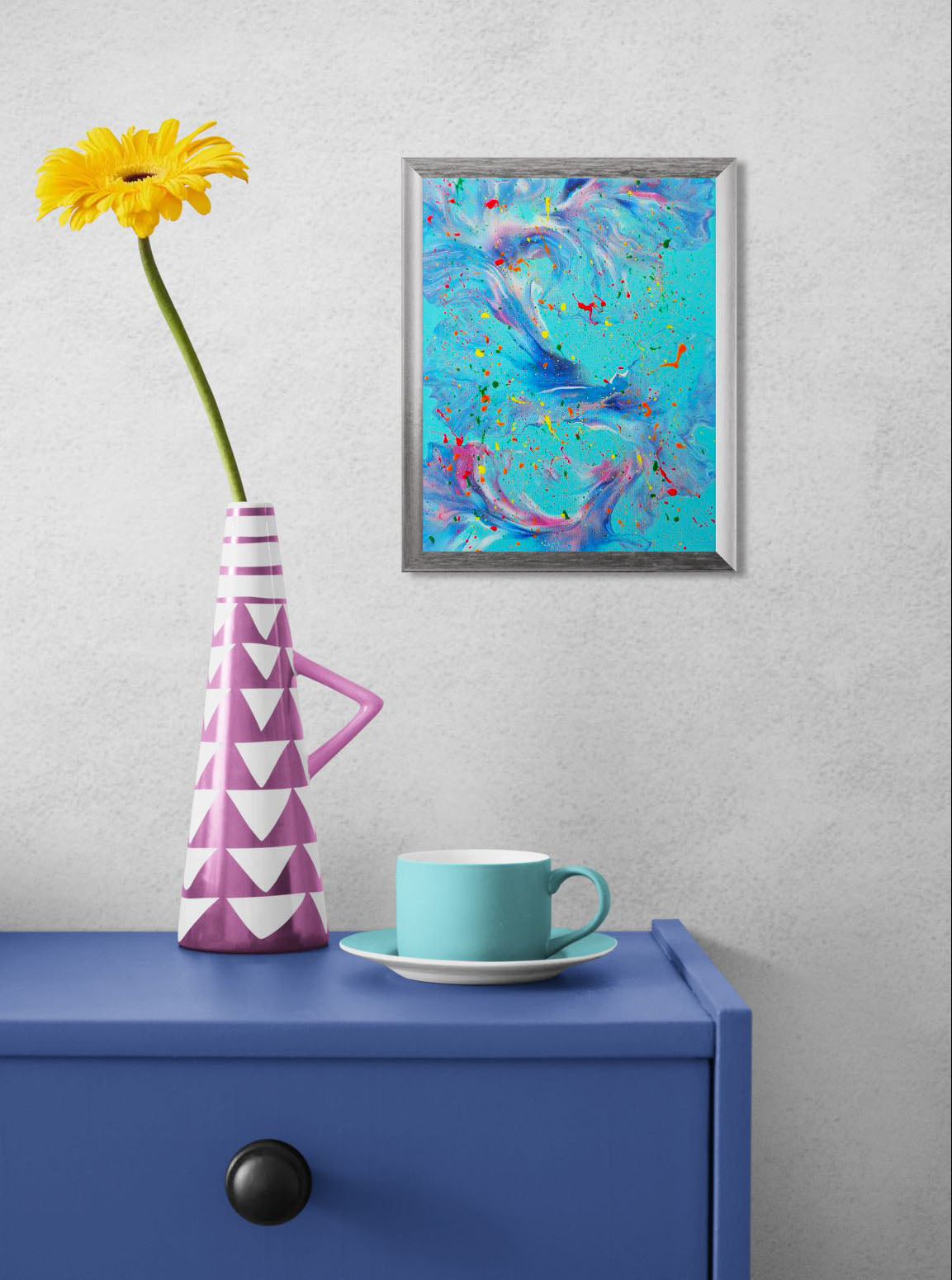 The painting hangs on a light grey wall above a bold blue table with a turquoise coffee cup and a pink geometric vase holding a single yellow flower. The artwork adds a vibrant, whimsical energy to the room.