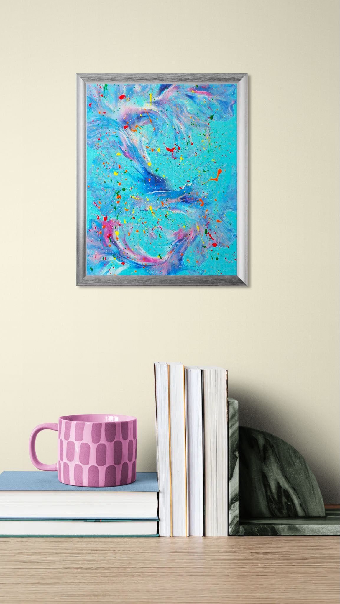The painting displayed in a cozy home office setting with soft neutral walls. It hangs above a stack of books and a pink-patterned mug, bringing a playful pop of color to the decor.